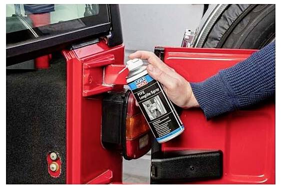 Eine Person sprüht Schmiermittel auf das Türscharnier einer roten Autotür zur Wartung, um einen reibungslosen Betrieb zu gewährleisten und Rostbildung zu verhindern.