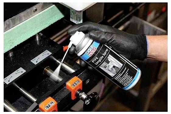 Eine behandschuhte Hand sprüht Schmiermittel aus einer Dose auf ein Metallteil einer Maschine, vermutlich zur Wartung oder zur Reibungsverringerung.