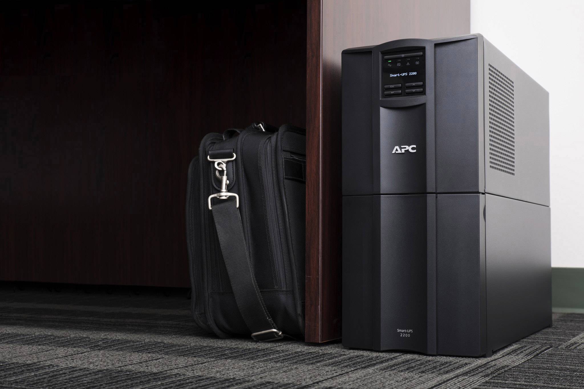Ein schwarzes APC-Backup-Gerät steht auf einem Teppichboden neben einer schwarzen Tasche, nahe einem Holztisch oder -schrank.