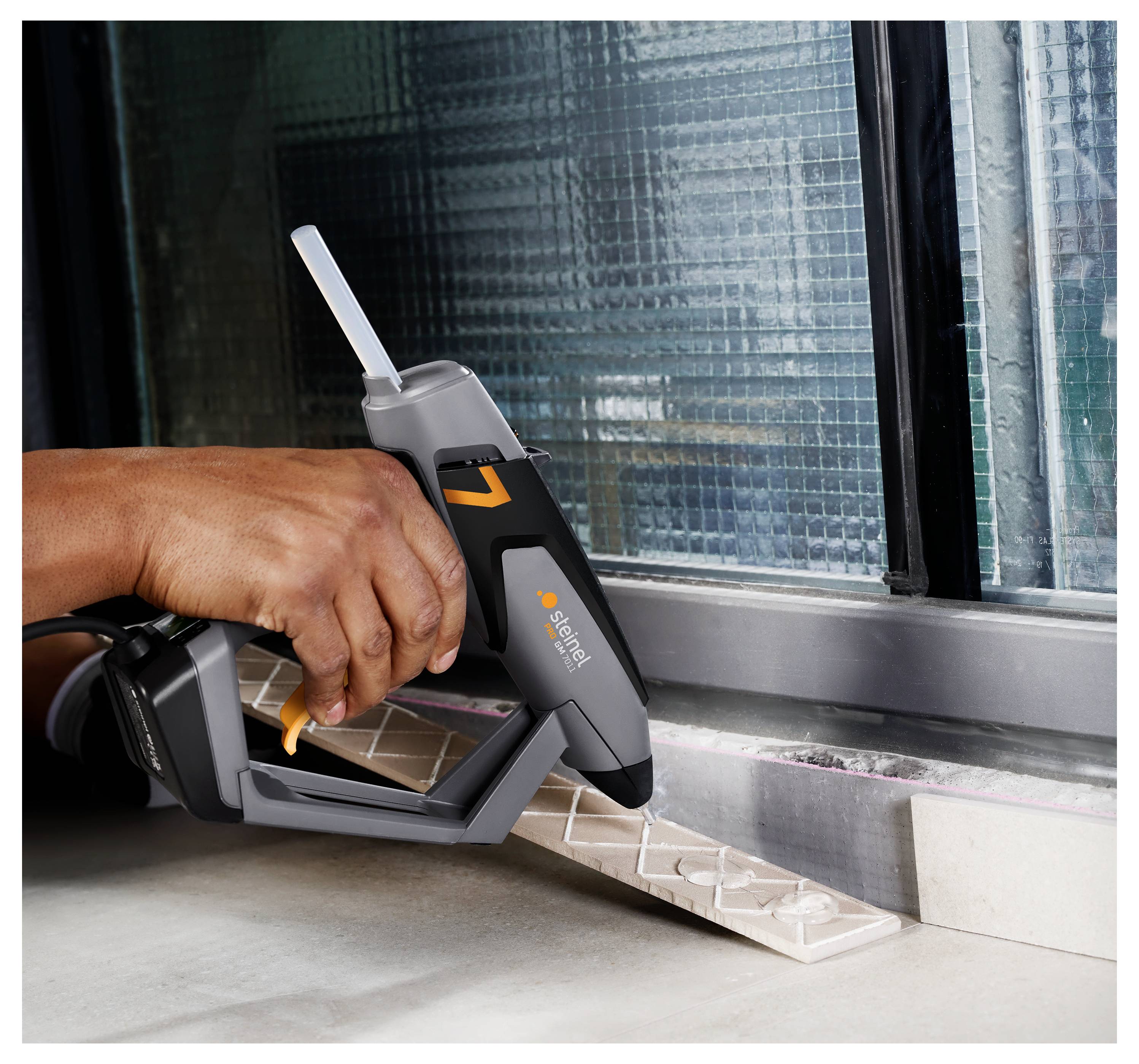 Hand mit einem kabellosen Applikator, der weißes Dichtungsmittel entlang der Kante eines Fensterrahmens auf einer Werkbank aufträgt.