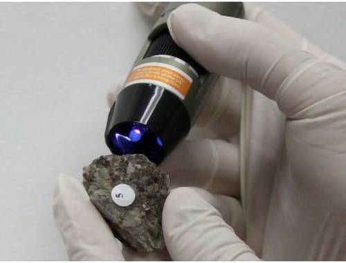 Behandschuhte Hände halten eine mit '5' beschriftete Gesteinsprobe, die mit einem leuchtenden wissenschaftlichen Gerät untersucht wird.