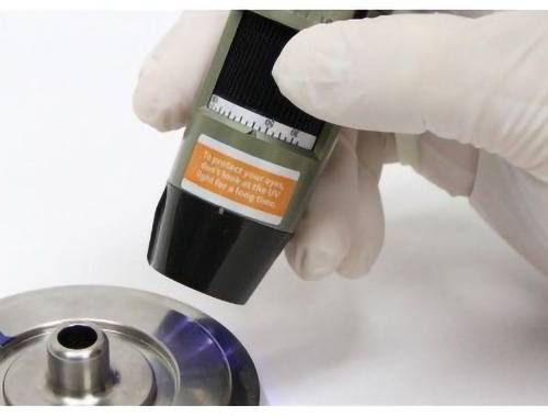 Eine behandschuhte Hand trägt Klebstoff aus einer Tube auf eine Metalloberfläche auf, wobei der Text auf der Tube vor UV-Lichtexposition zur effektiven Verklebung warnt.