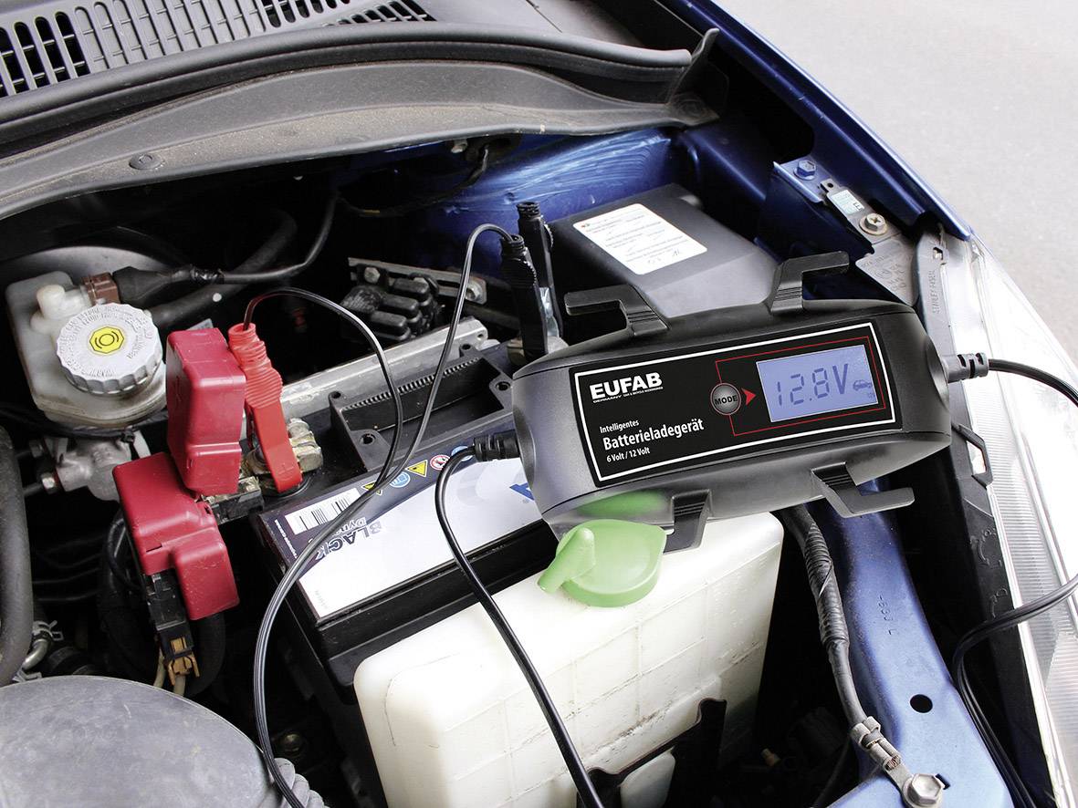 Ein Batterieladegerät mit digitaler Anzeige zeigt 12,8V, angeschlossen an eine Autobatterie in einem Fahrzeugmotorraum.