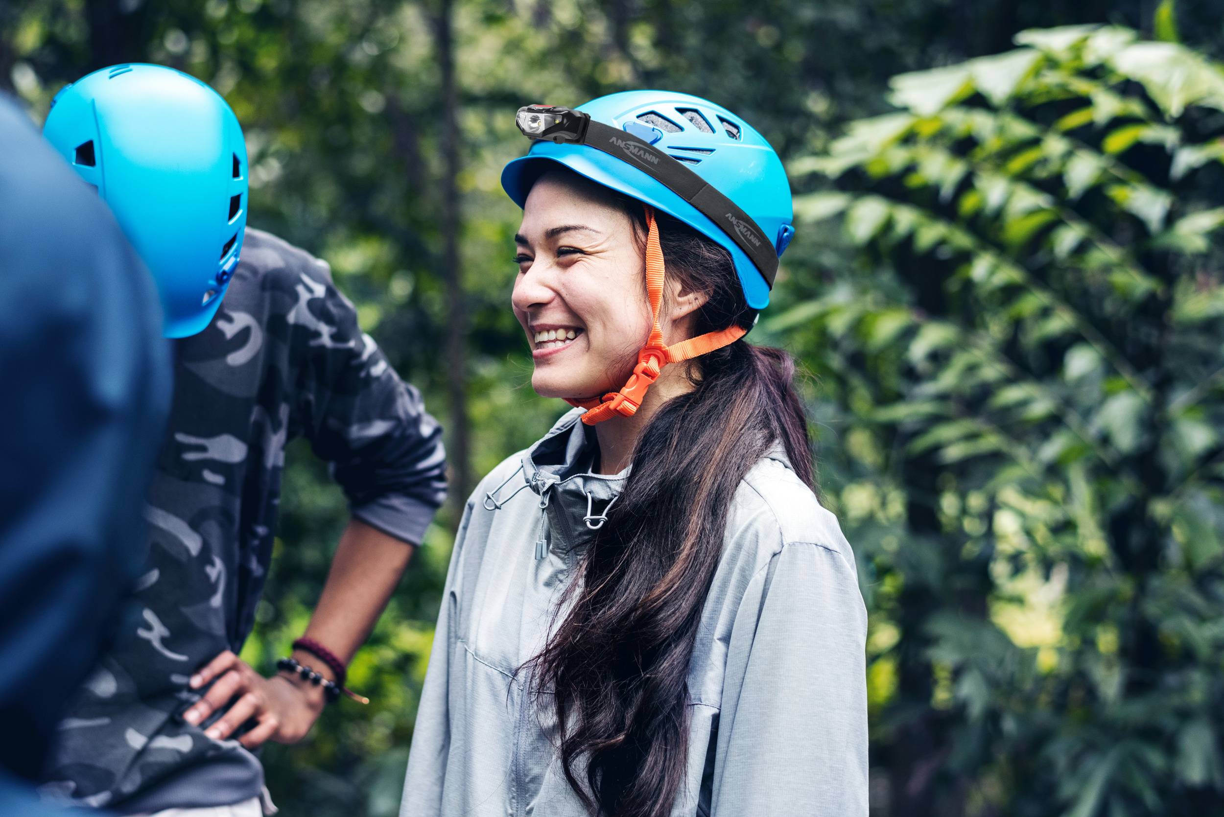 Eine Person lächelt im Wald und trägt einen blauen Helm. Umgeben von weiteren Personen und üppigem Grün.