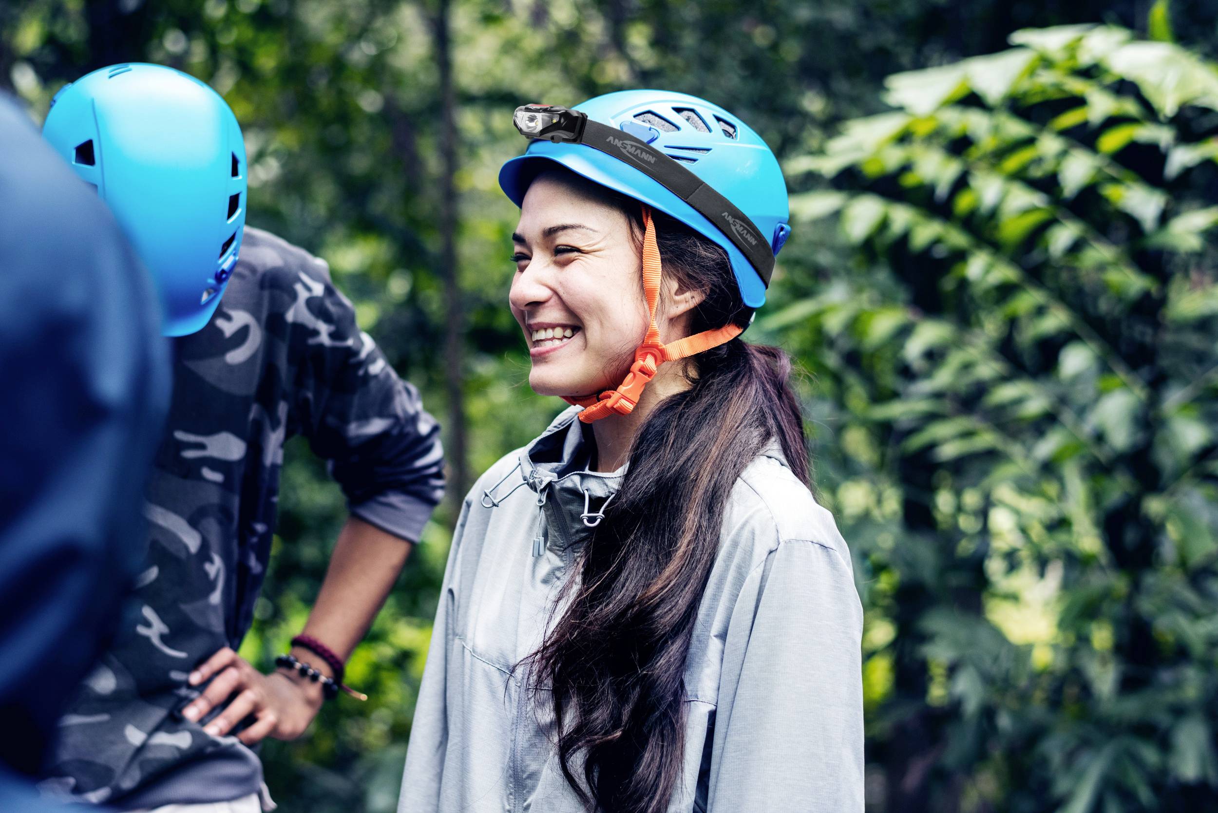 Eine lächelnde Person trägt einen blauen Kletterhelm und steht in einem bewaldeten Gebiet, umgeben von grüner Vegetation.