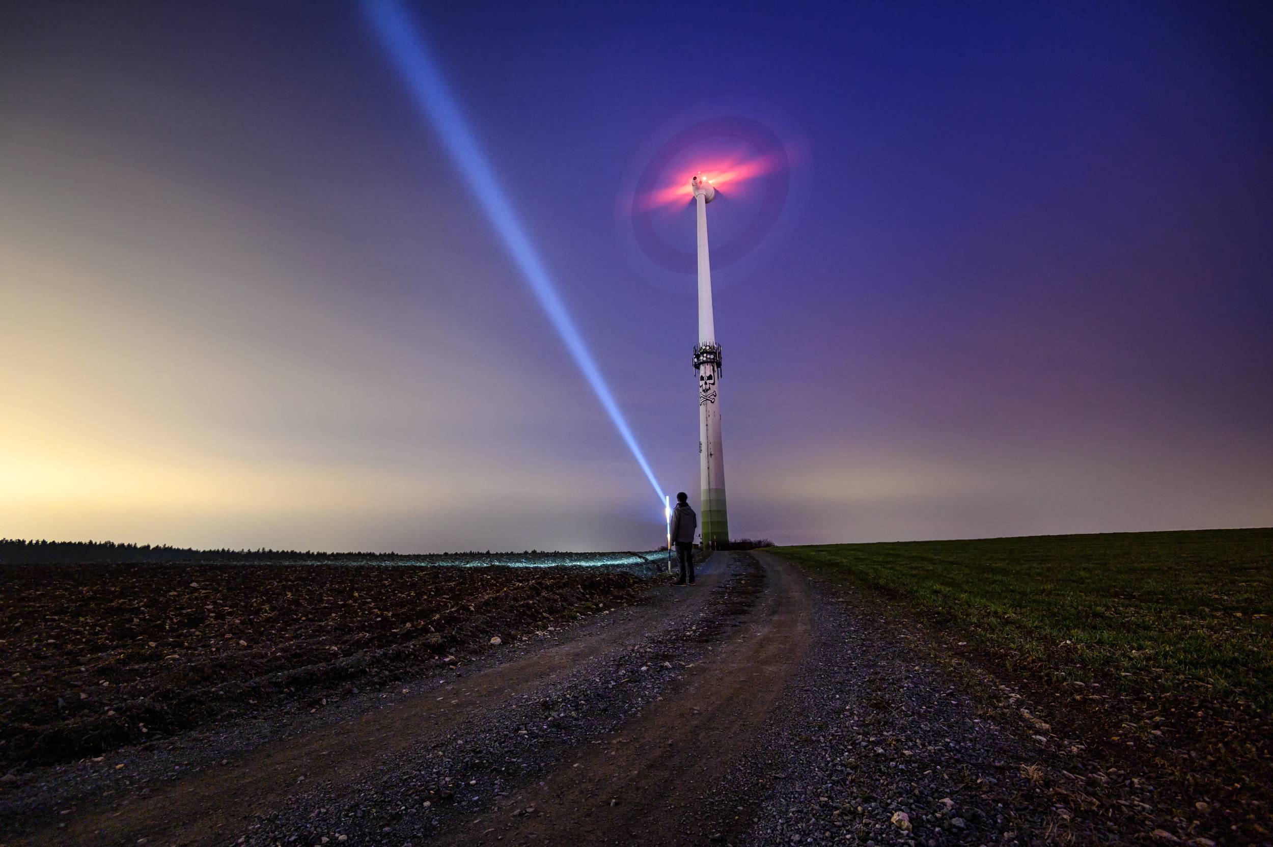 Eine Person steht auf einem Feldweg bei Nacht und leuchtet mit einer Taschenlampe auf ein beleuchtetes Windrad unter einem farbenfrohen Himmel.