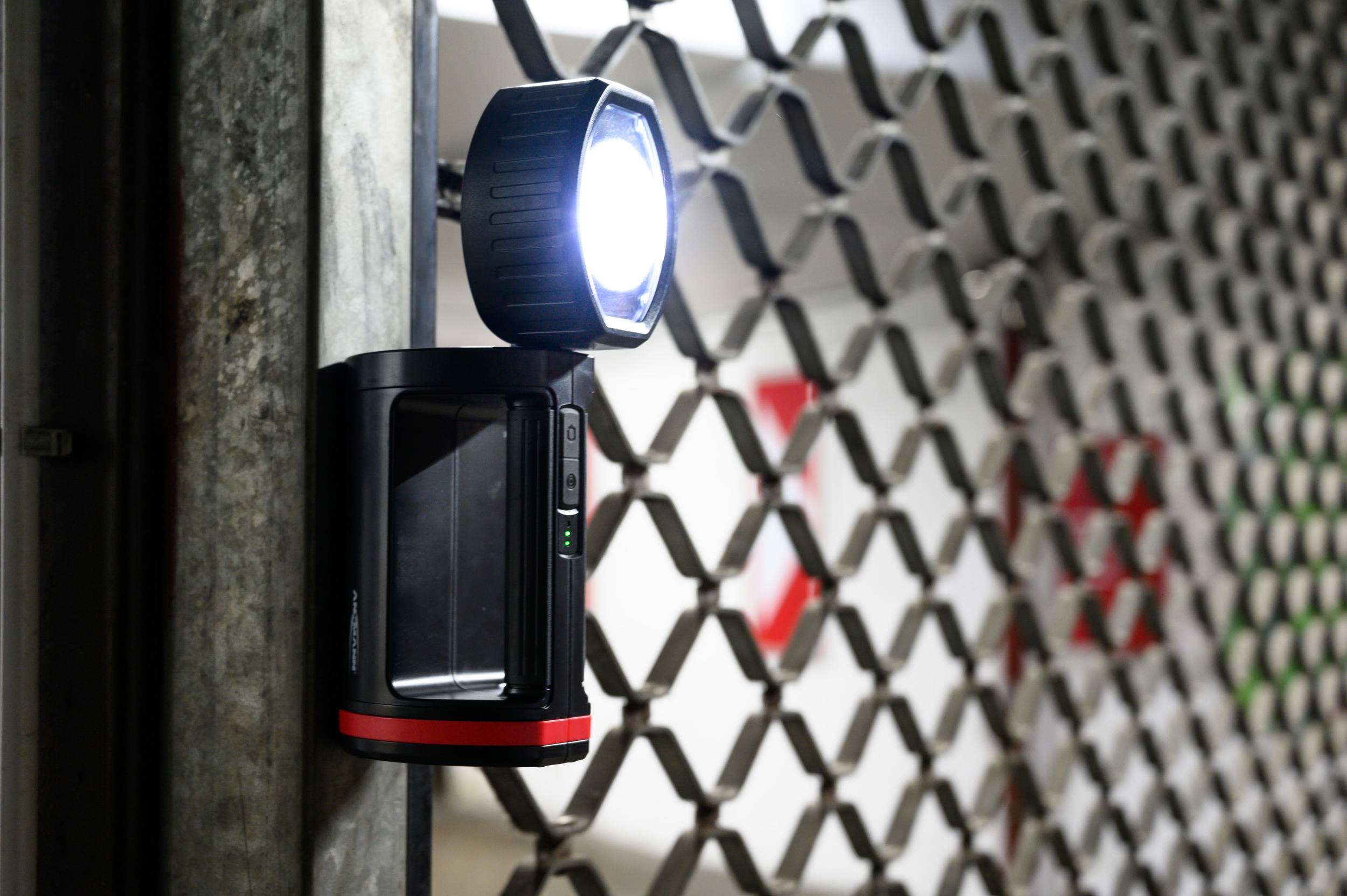 Eine Überwachungskamera mit hellem Licht ist an einem Metallzaun befestigt. Im Hintergrund ist teilweise ein rotes Schild zu sehen.