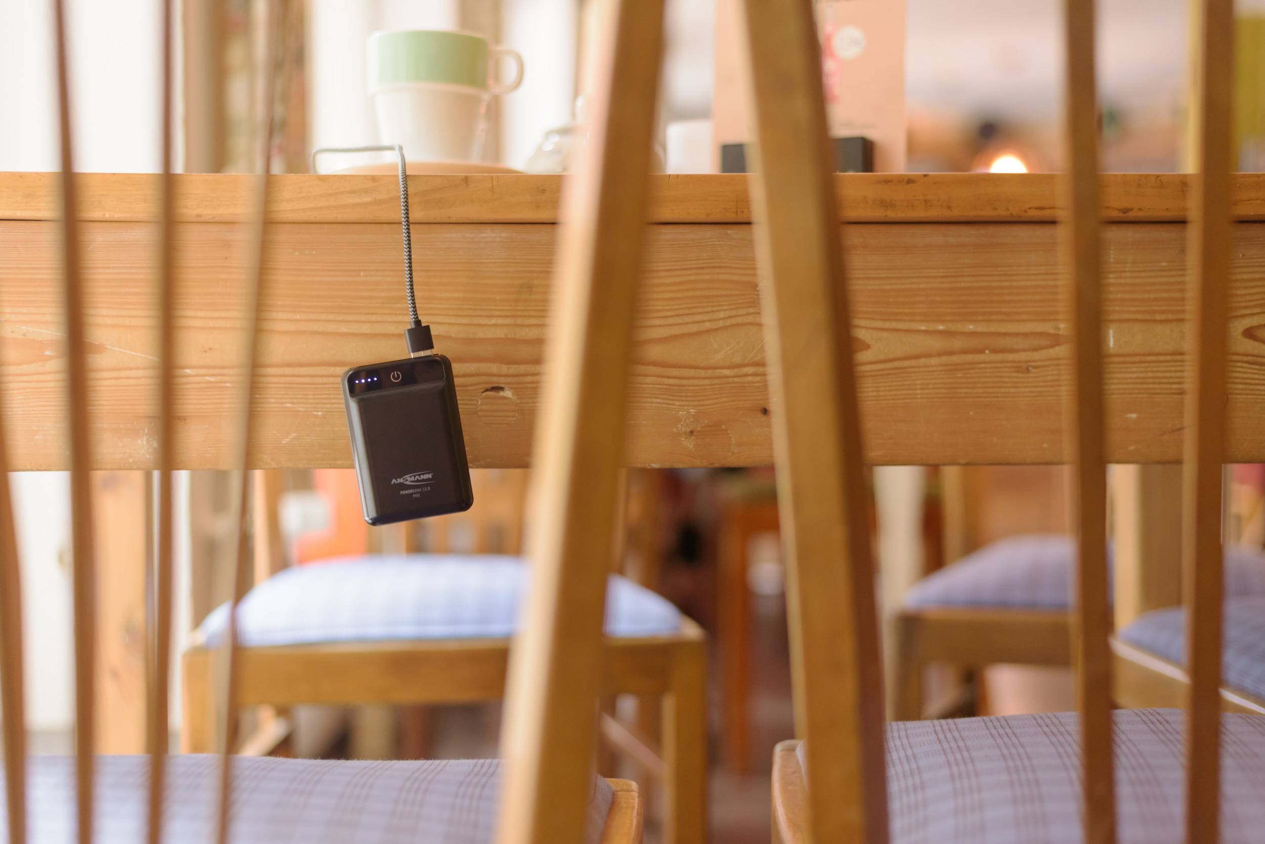 Ein Smartphone hängt umgedreht an einem Ladekabel unter einem Holztisch in einem gemütlichen Raum mit Stühlen und Tassen.