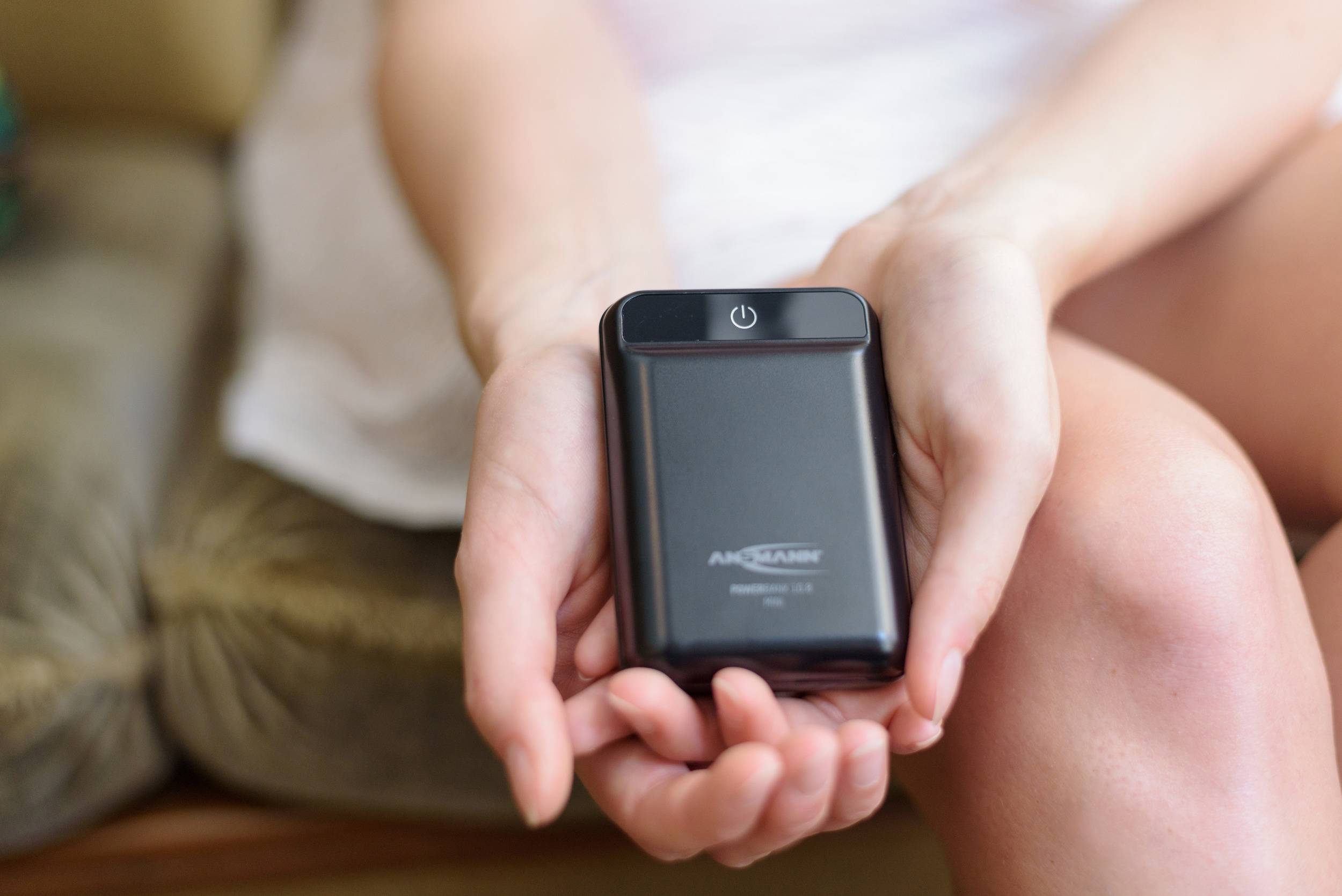 Person hält eine kleine, schwarze Powerbank in beiden Händen. Power-Taste oben sichtbar, auf einem Sofasitz sitzend.
