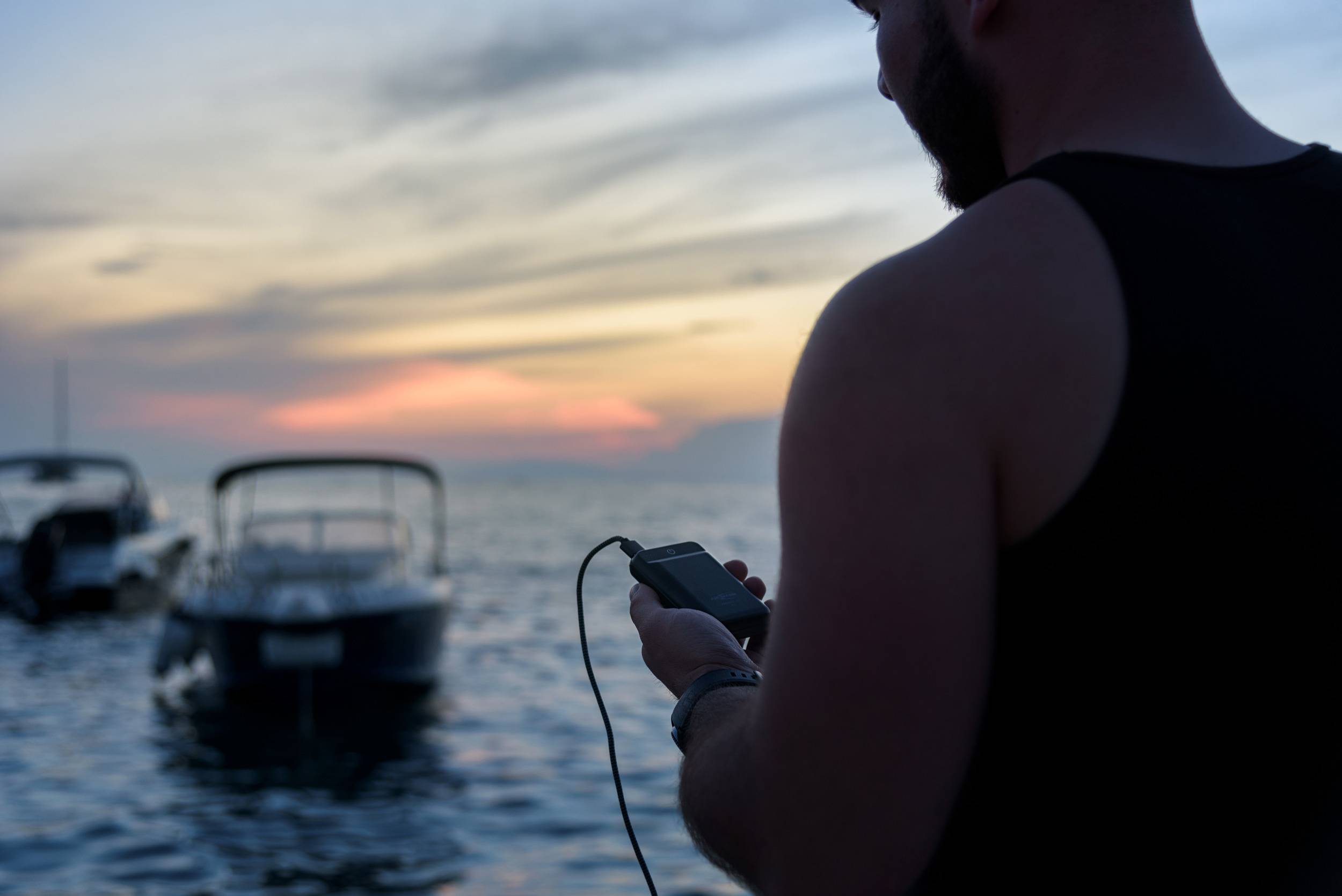 Eine Person hält ein Smartphone in der Hand und steht am Ufer mit Booten im Wasser. Der Himmel zeigt ein farbenfrohes Abendrot.