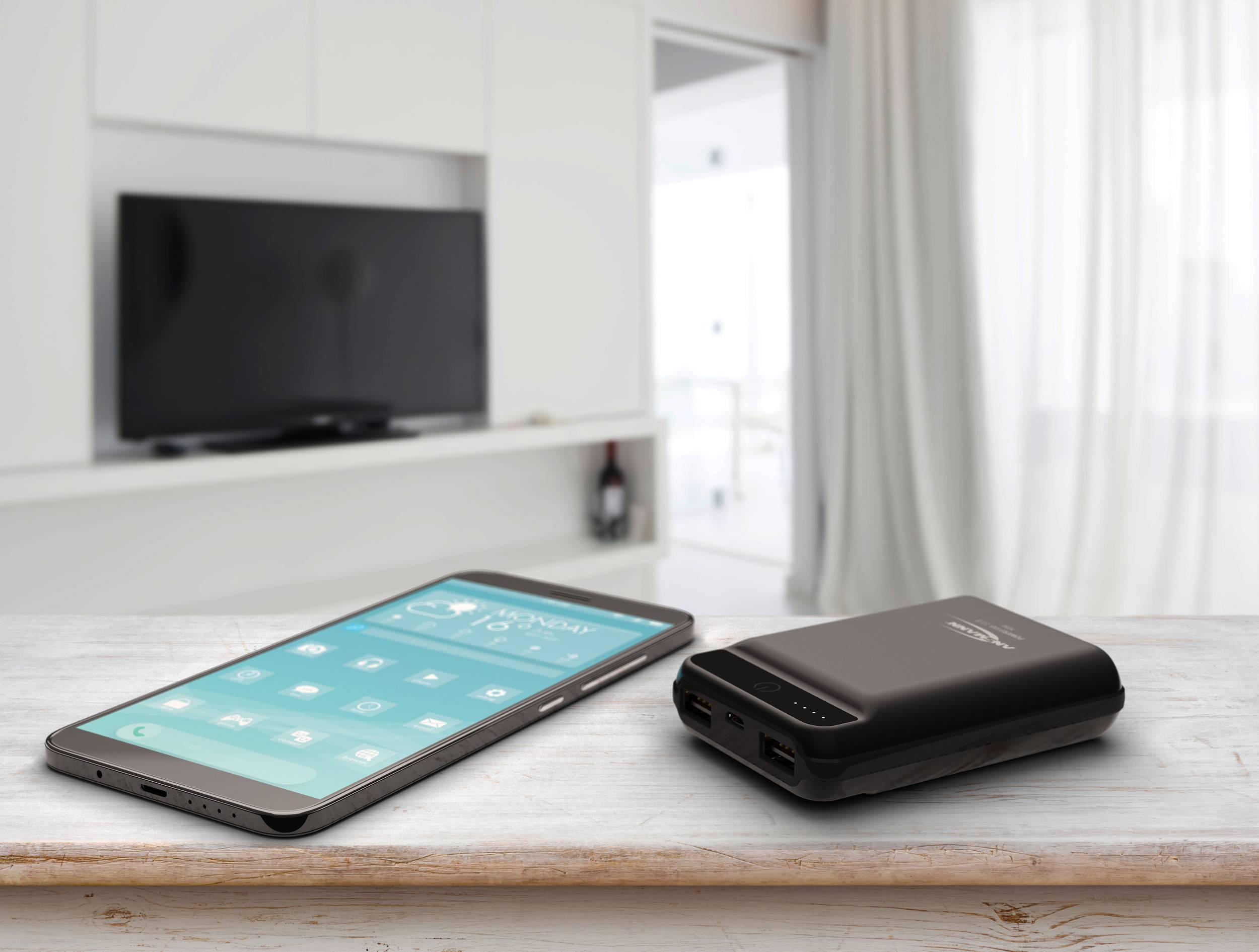 Ein Smartphone und eine Powerbank liegen auf einem Holztisch in einem modernen Wohnzimmer. Im Hintergrund ist ein Fernseher zu sehen.