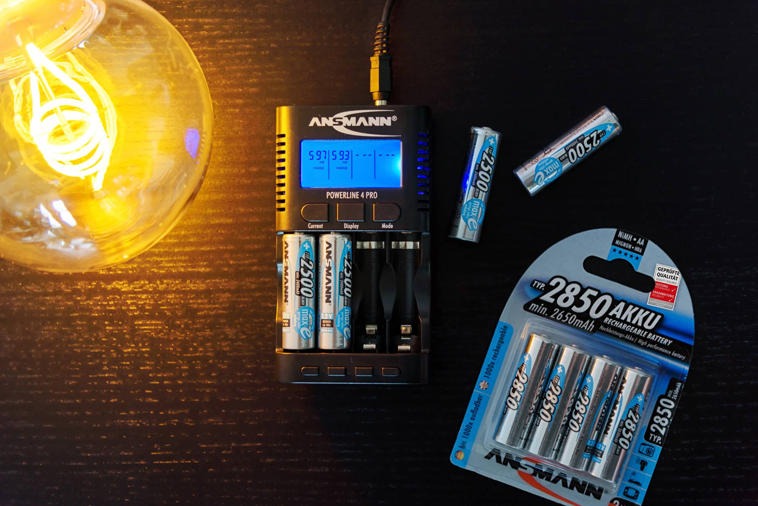 Ein Ladengerät lädt AA-Batterien neben einer geöffneten Verpackung und ein leuchtendes LED-Licht erhellt die Szene auf dunklem Tisch.