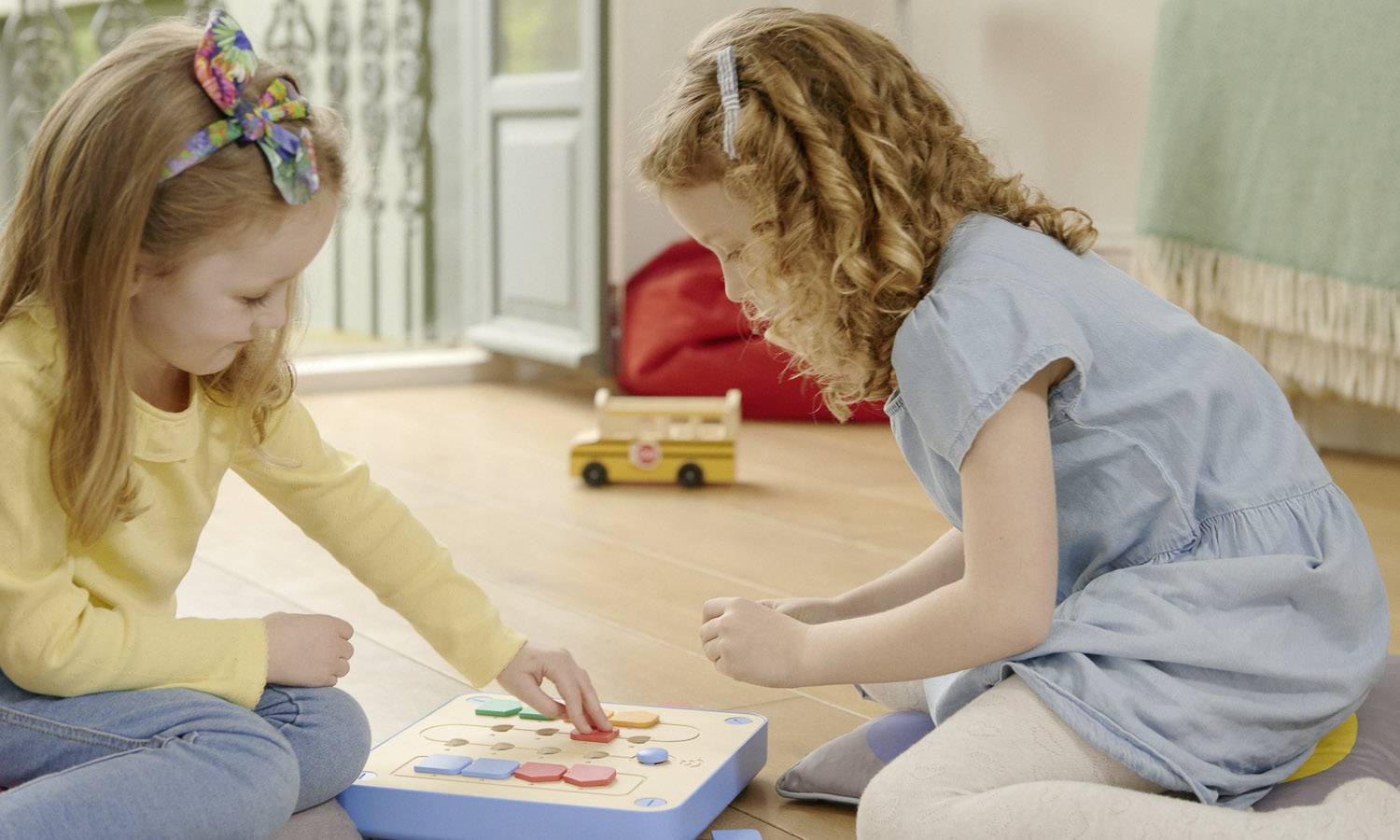 Zwei Mädchen spielen auf dem Fußboden mit einem Holzpuzzle. Sie zeigen Konzentration und Freude in einem hellen, gemütlichen Raum.