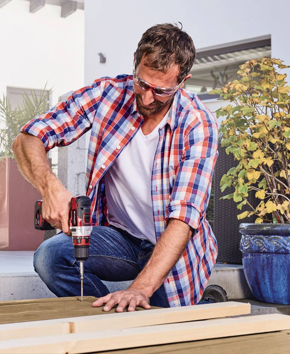 Ein Mann im Karomusterhemd benutzt eine Bohrmaschine, um Holz auf einer Terrasse zu bearbeiten. Im Hintergrund sind Pflanzen sichtbar.