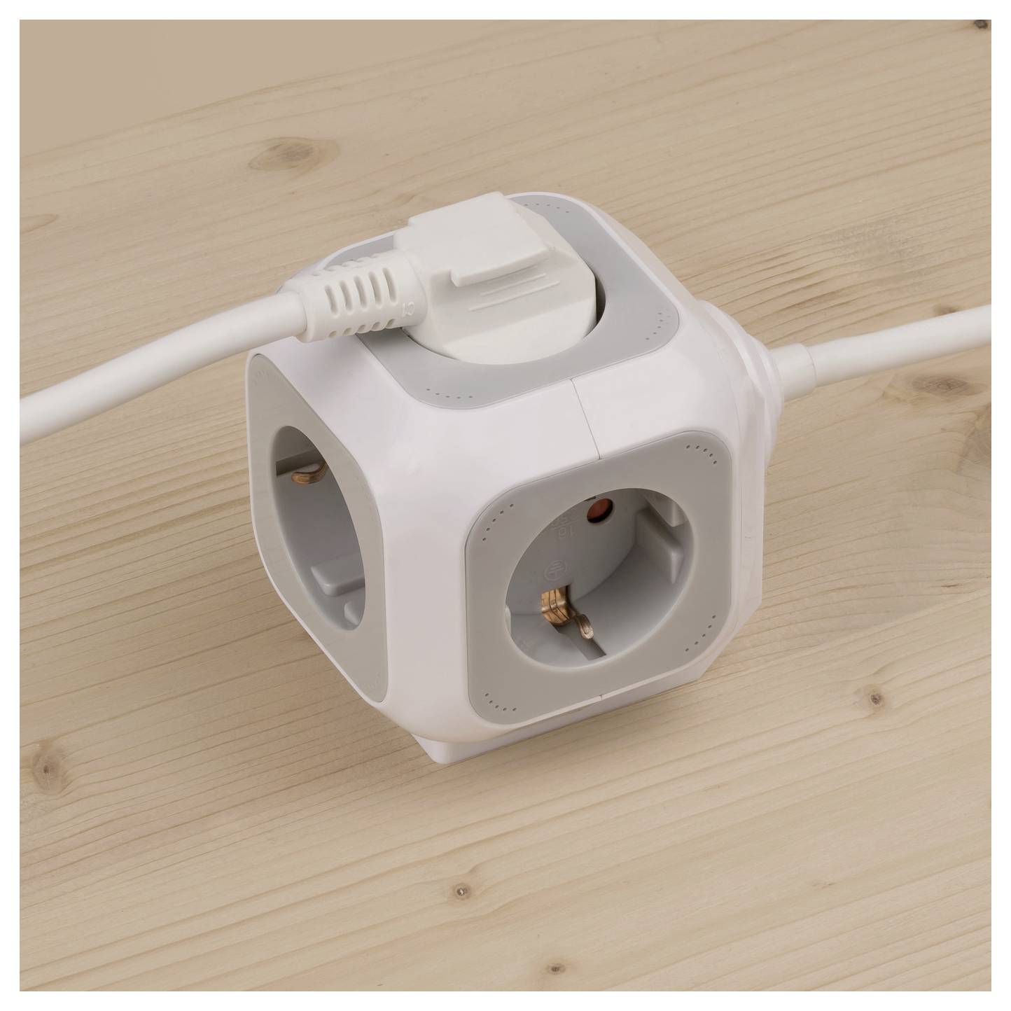 Weiße würfelförmige Steckdosenleiste mit mehreren Steckdosen auf einer Holzoberfläche, mit einem eingesteckten Stecker oben und Kabeln, die auf beiden Seiten herausragen.