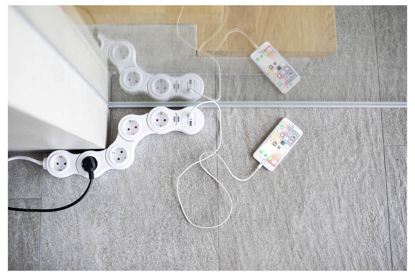 Zwei Smartphones, die durch weiße Ladekabel verbunden sind, liegen auf einem Teppich neben einer verspiegelten Wand und Steckerleisten, die in eine Steckdose eingesteckt sind.