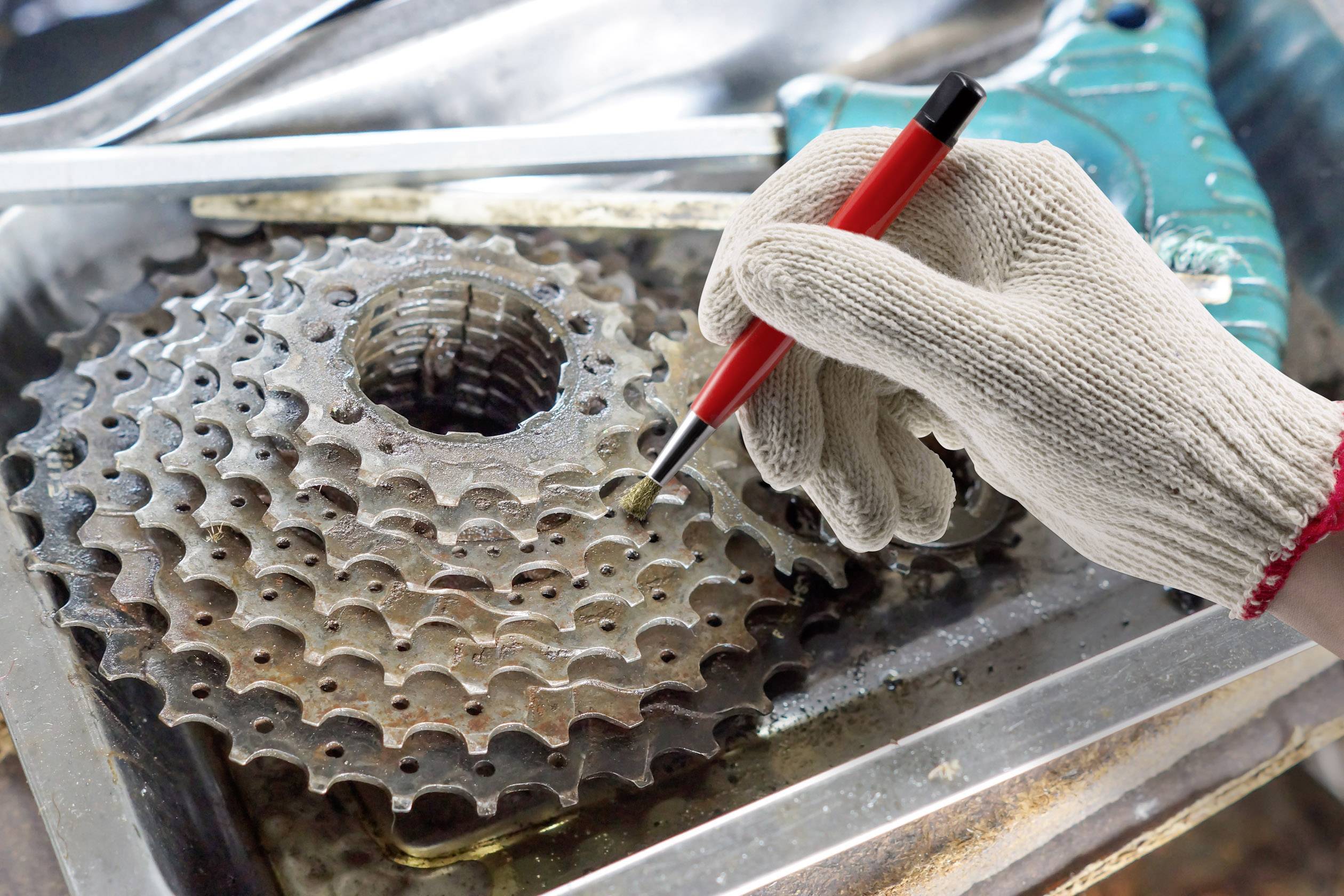 Hand mit Handschuh reinigt Fahrradkassette mit rotem Werkzeug. Hintergrund aus Metallteilen, Arbeit in Fahrradwerkstatt.