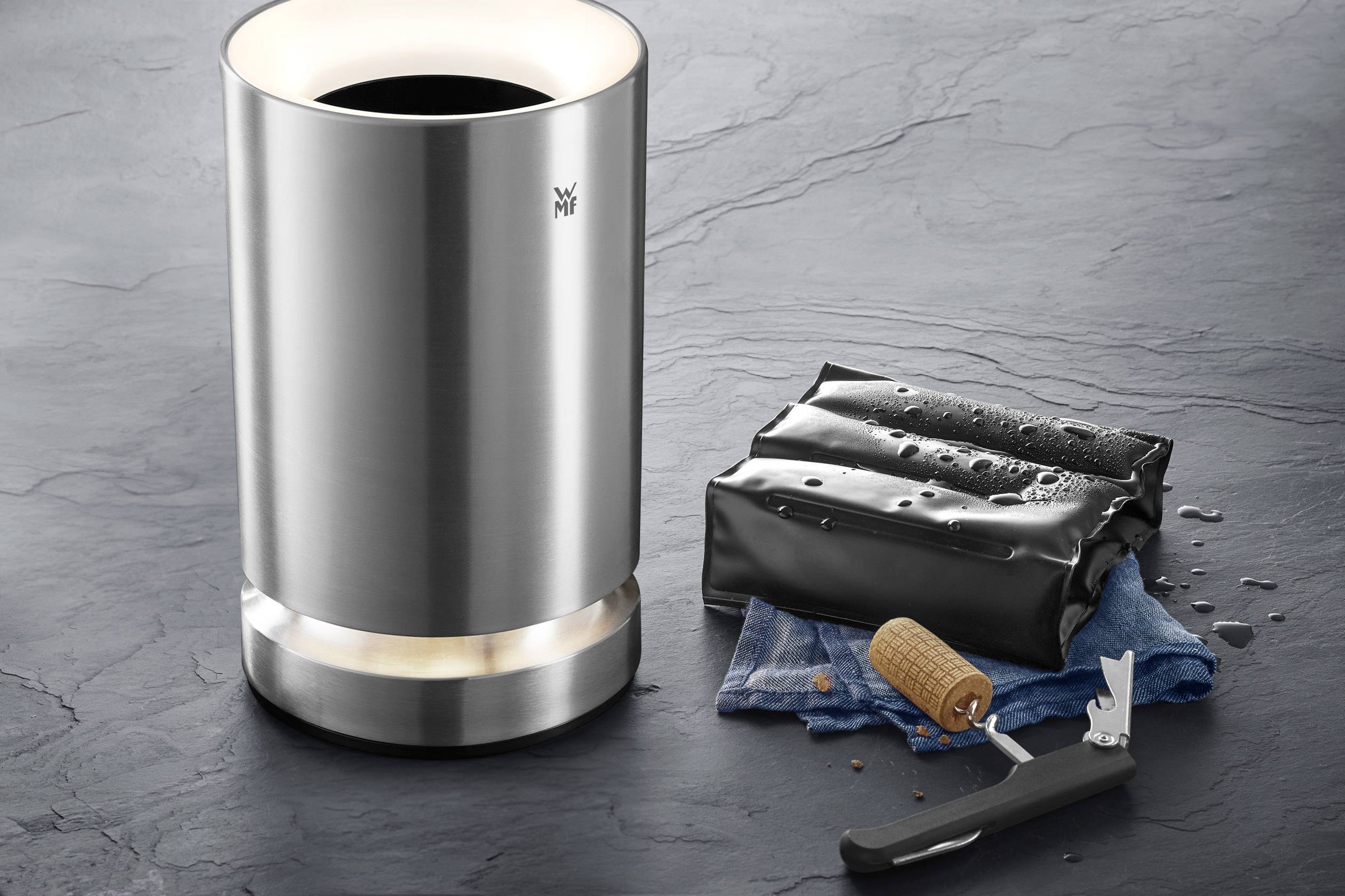 Ein elektrischer Flaschenkühler aus Edelstahl neben einem geöffneten Flaschenverschluss auf einem blauen Tuch.