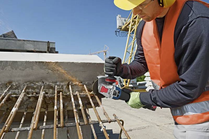 Ein Bauarbeiter in Schutzausrüstung verwendet eine Winkelschleifmaschine, um rostige Stahlbewehrungsstäbe zu durchtrennen, die aus Beton auf einer Baustelle herausragen.