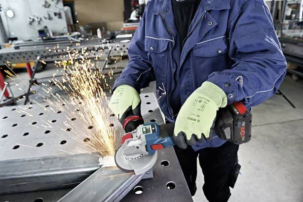 Eine Person in Arbeitskleidung schleift mit einem Winkelschleifer Metall, Funken fliegen. Sie trägt grüne Handschuhe und eine blaue Jacke.