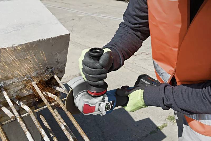 Person in Sicherheitsweste nutzt einen Winkelschleifer, um Metallstäbe aus Beton zu schneiden. Funken sprühen bei der Arbeit.