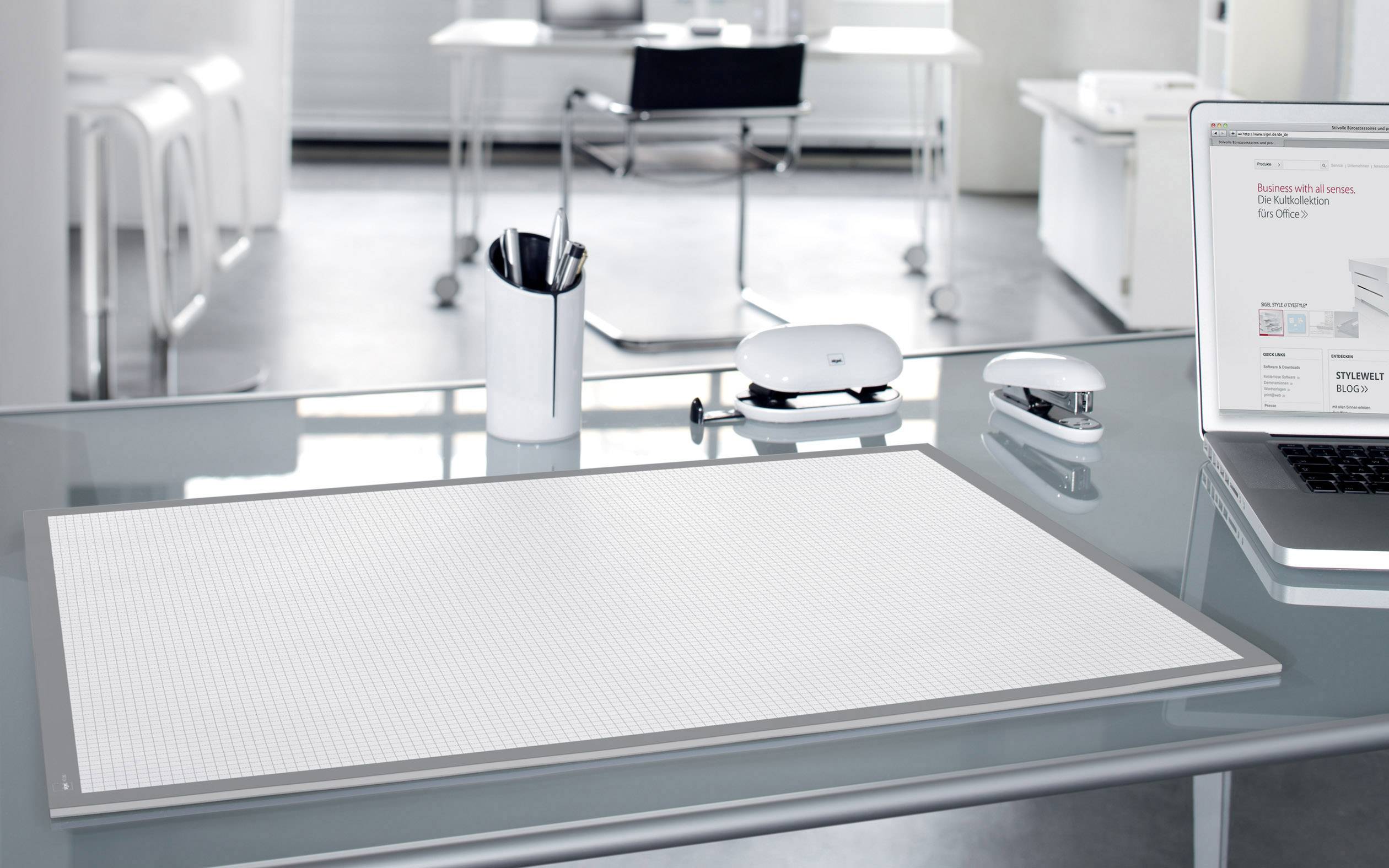 Moderner Arbeitsplatz mit transparentem Glastisch, darauf ein Laptop, Schreibtischunterlage, Stiftehalter, Locher und Maus. Helle, minimalistische Umgebung.
