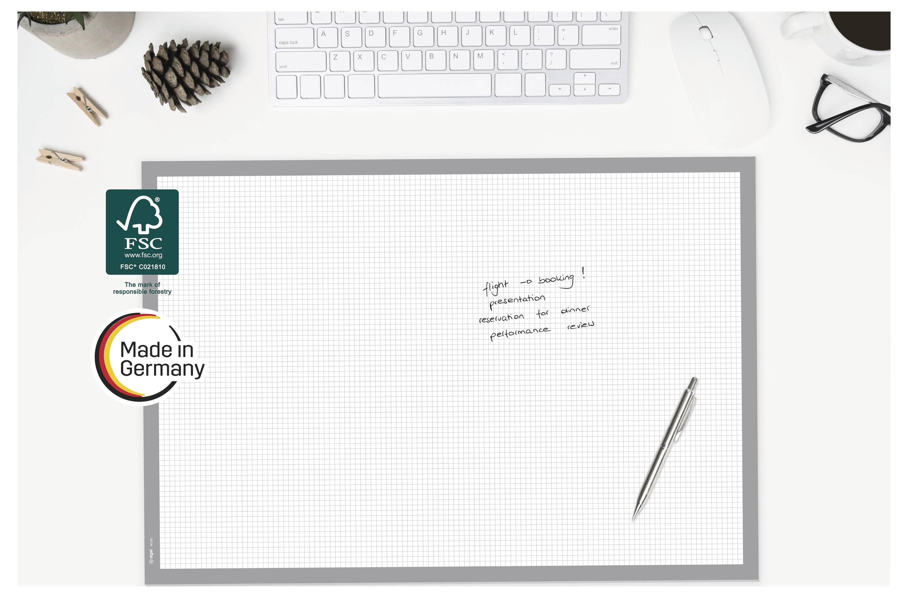Schreibtischszene von oben mit leerem kariertem Notizblock, Stift, Tastatur, Brille und FSC- sowie „Made in Germany"-Plaketten auf der linken Seite.
