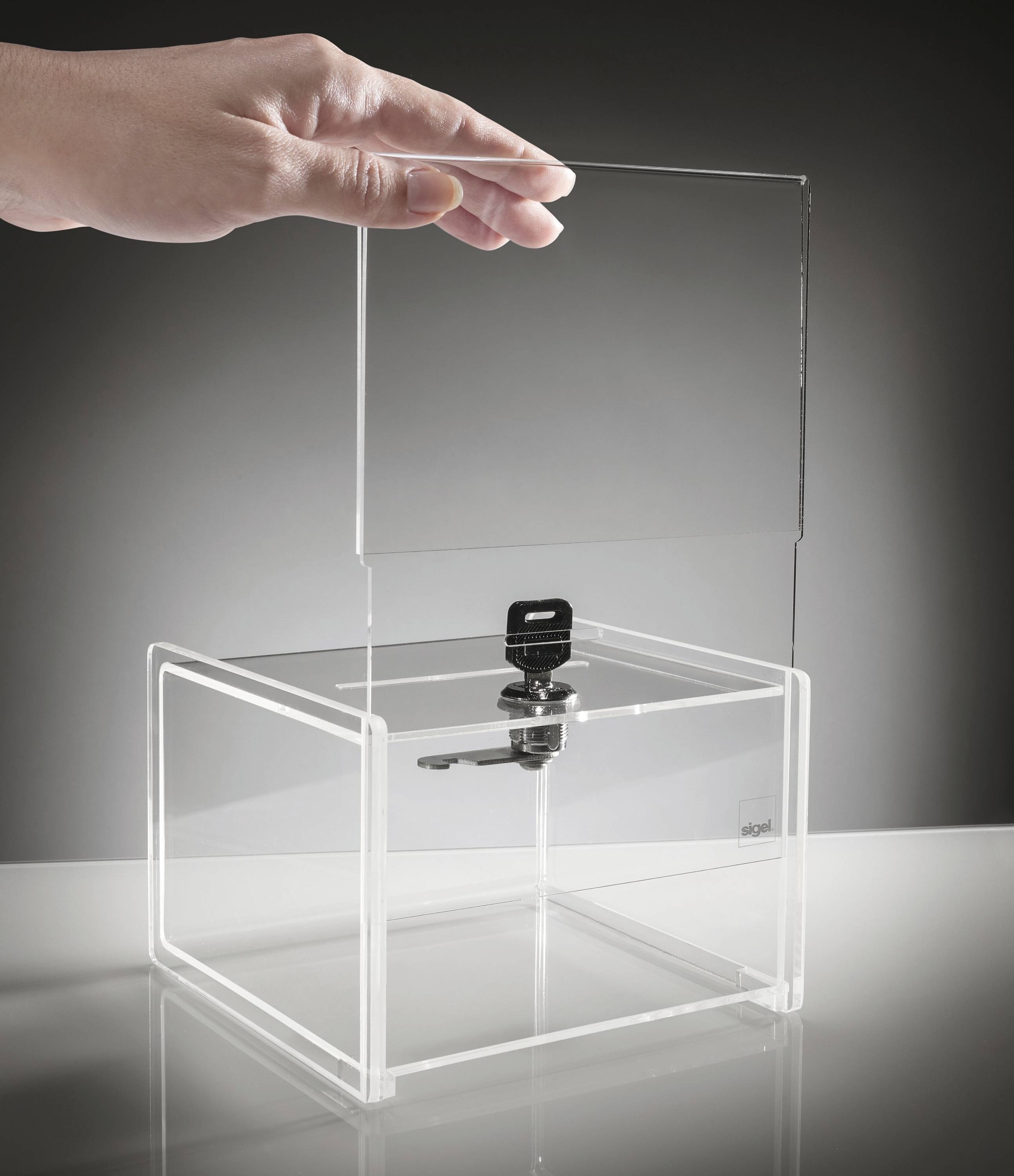 Eine Hand legt einen transparenten Acryldeckel auf eine durchsichtige Box mit einem Schloss, was Sicherheit oder den Schutz des Inhalts andeutet.