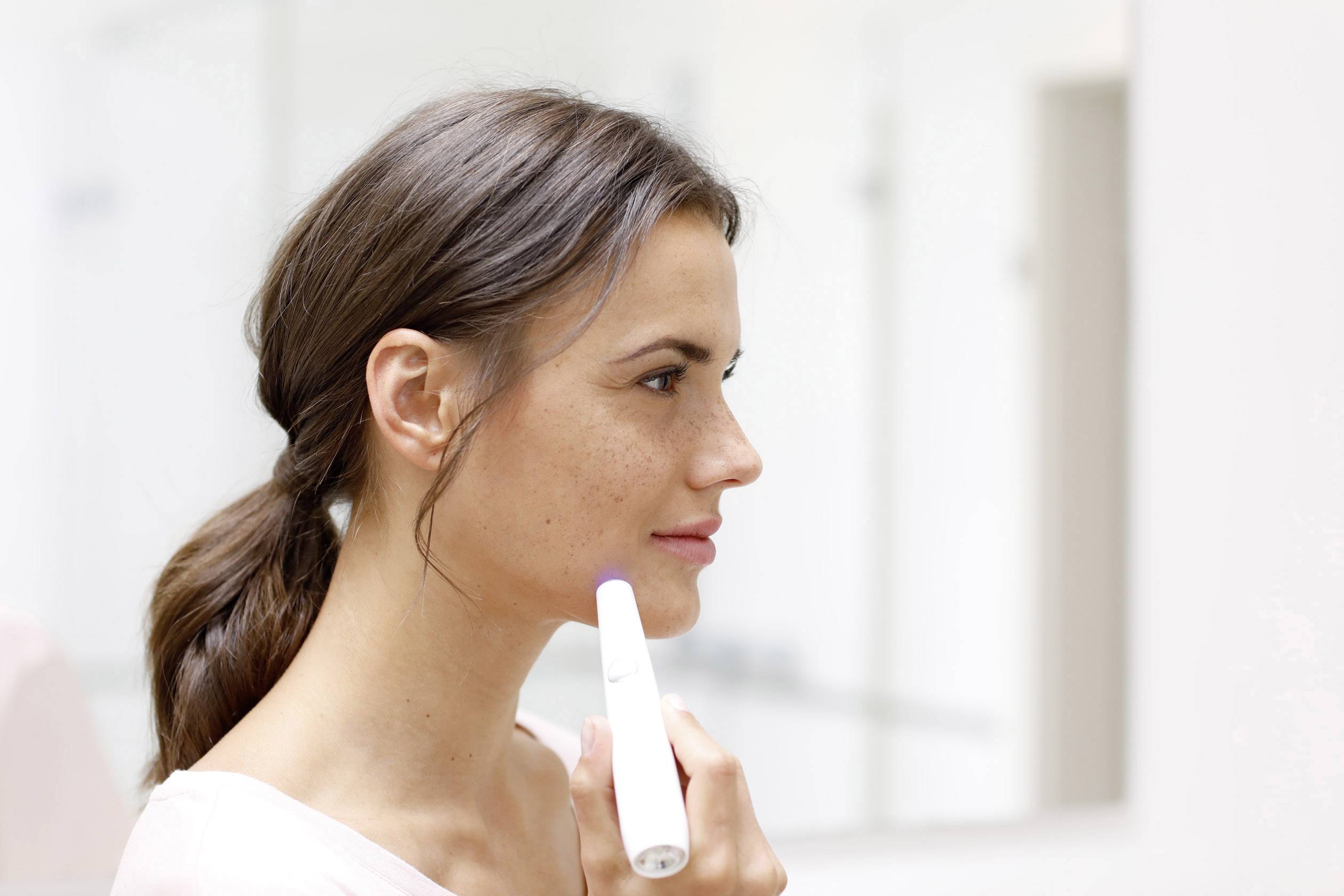 Frau mit Zopf hält einen Hautpflegestift an die Wange, Spiegel-Hintergrund.