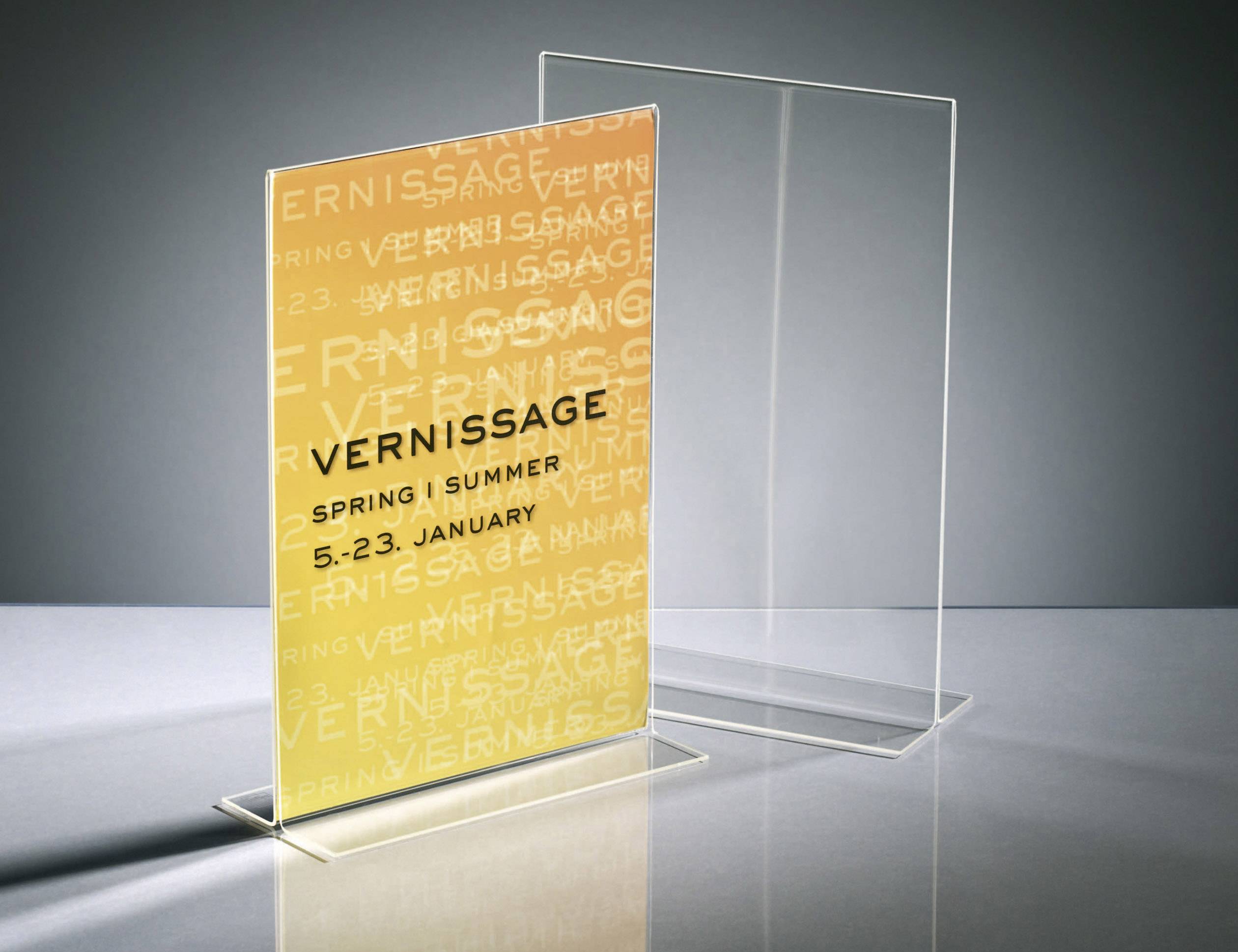 Ein Schild mit der Aufschrift 'Vernissage Spring | Summer 5.-23. January' auf orangefarbenem Hintergrund, steht in einem transparenten Ständer.