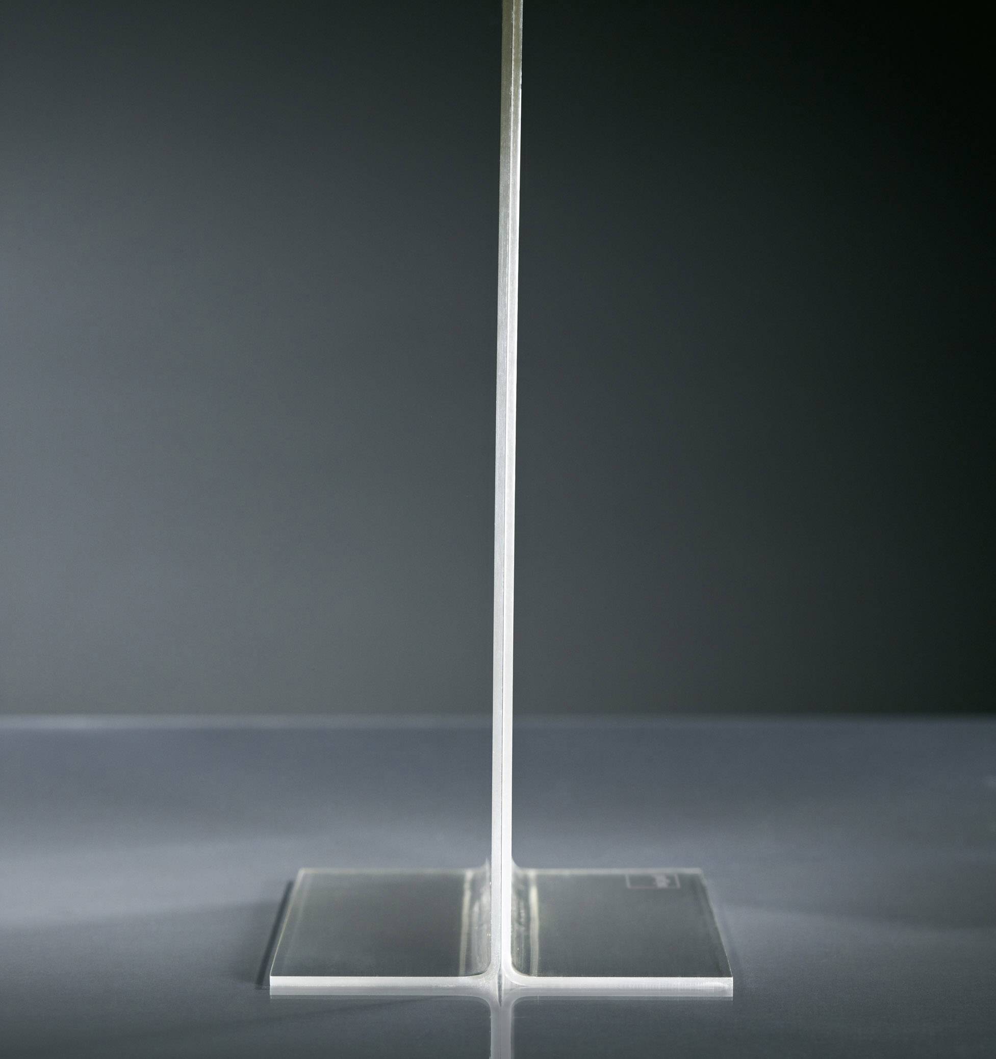 Klares Acrylobjekt in T-Form vor grauem Hintergrund, auf einer spiegelnden Oberfläche stehend.