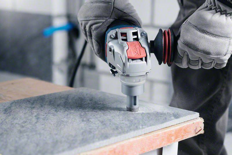 Ein Arbeiter bohrt mit einer Bohrmaschine ein Loch in eine graue Steinplatte, trägt dabei Schutzhandschuhe und Arbeitskleidung.