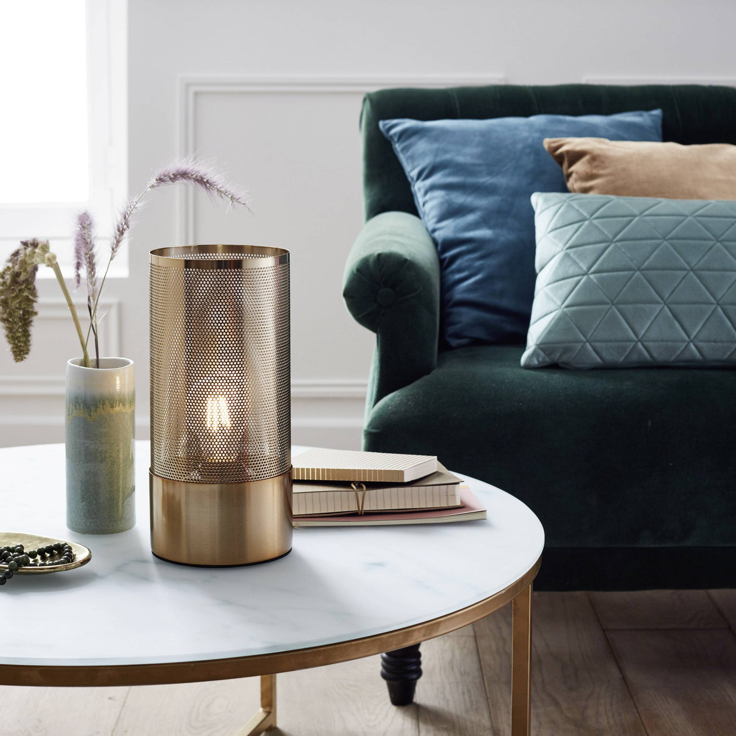 Ein Tisch mit einer Lampe aus Metallgeflecht, daneben ein Vase mit Gräsern und ein Stapel Bücher, vor einem grünen Sofa mit bunten Kissen.