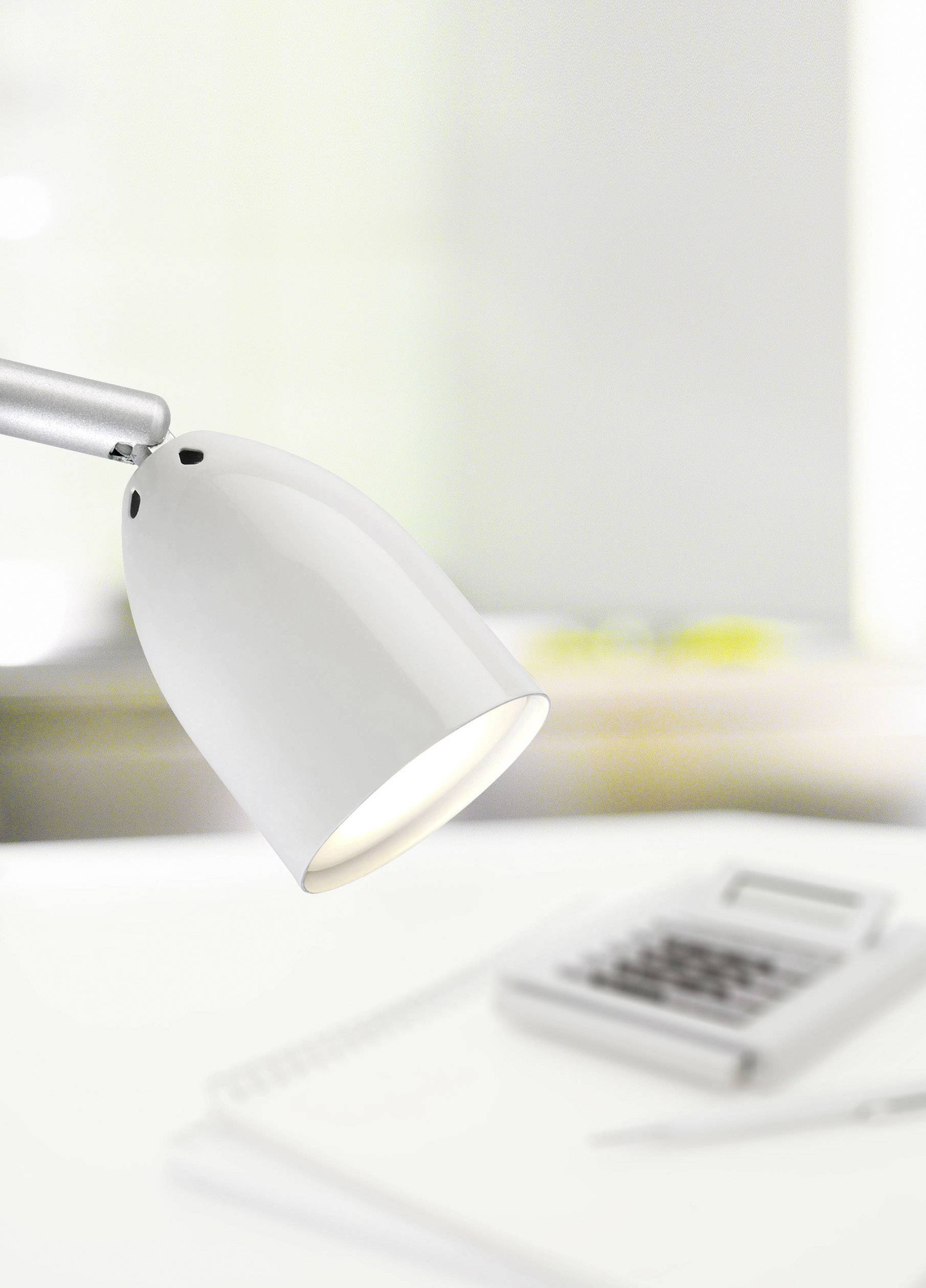 Eine weiße Schreibtischlampe beleuchtet ein Notizbuch und einen Taschenrechner auf einem Schreibtisch, die Umgebung ist unscharf.