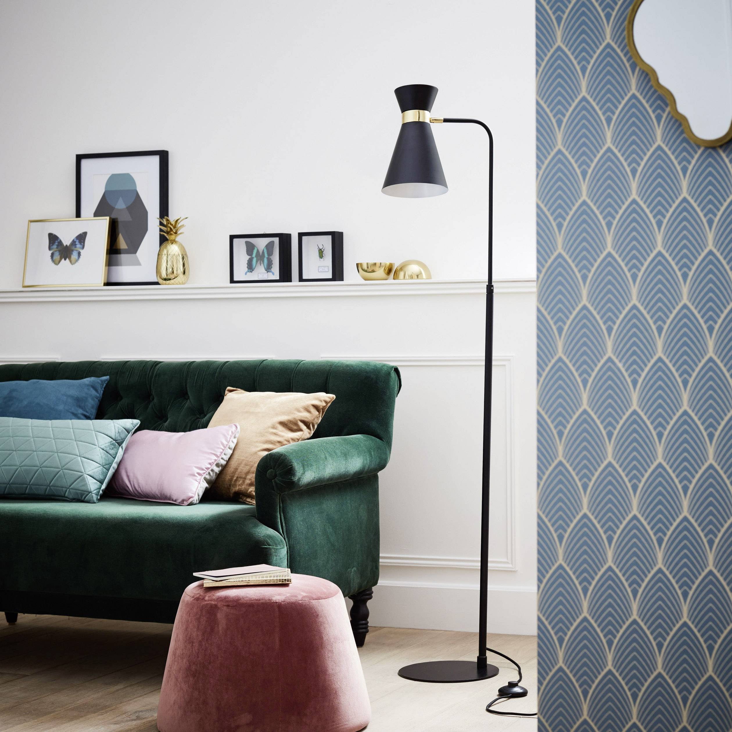 Ein stilvoll eingerichtetes Wohnzimmer mit einem grünen Sofa, einem pinken Hocker und einer schwarzen Stehlampe vor einer gemusterten Tapete.