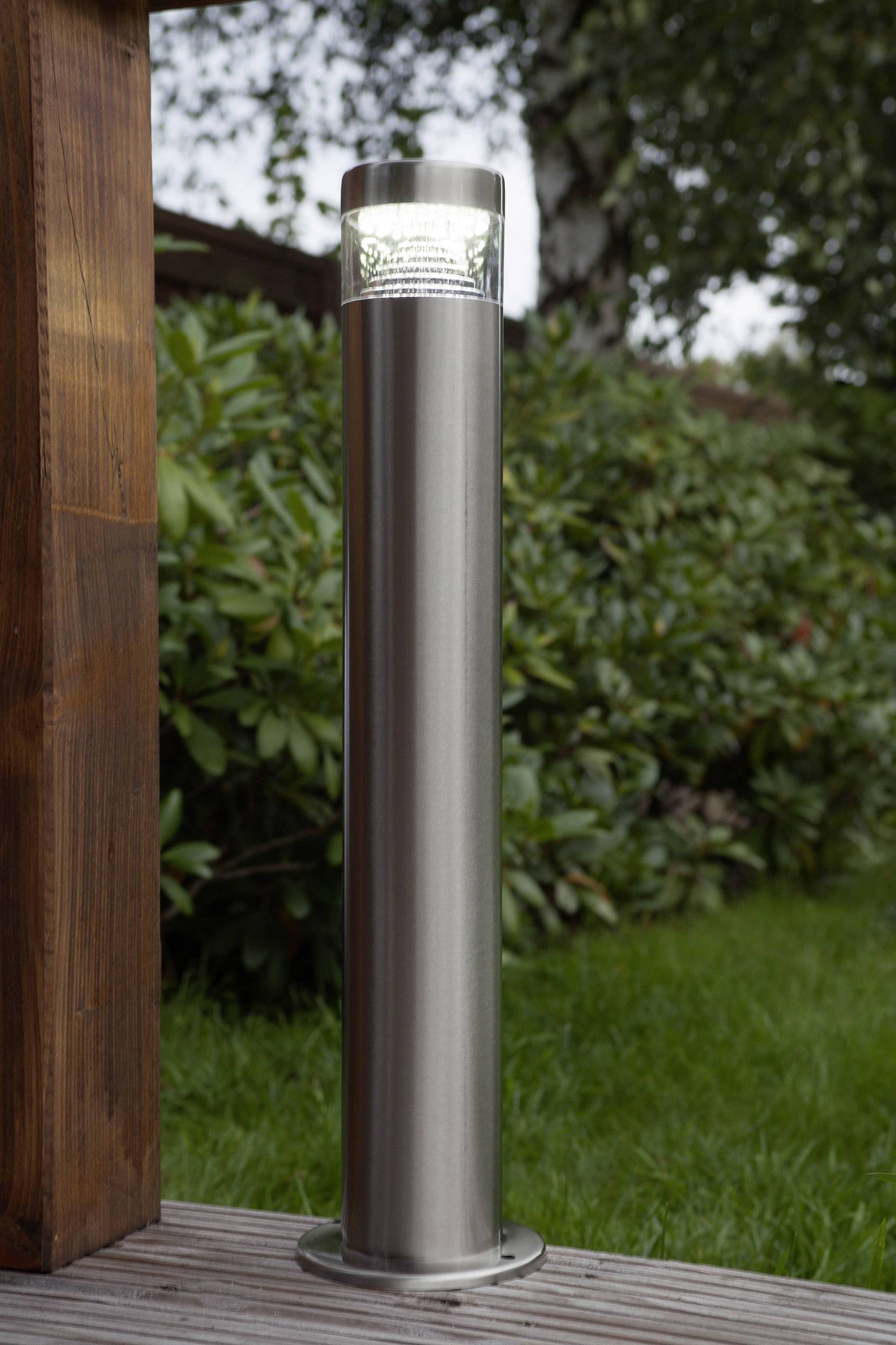 Edelstahl-Solarleuchte steht auf Holzterrasse, von Pflanzen umgeben.