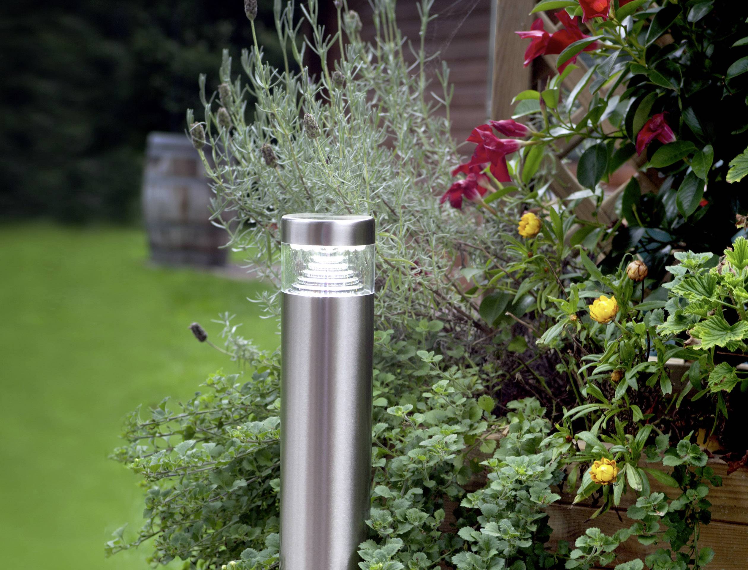 Eine moderne Gartenlampe aus Edelstahl steht inmitten üppiger grüner Pflanzen und bunten Blumen, die eine gemütliche Atmosphäre schaffen.