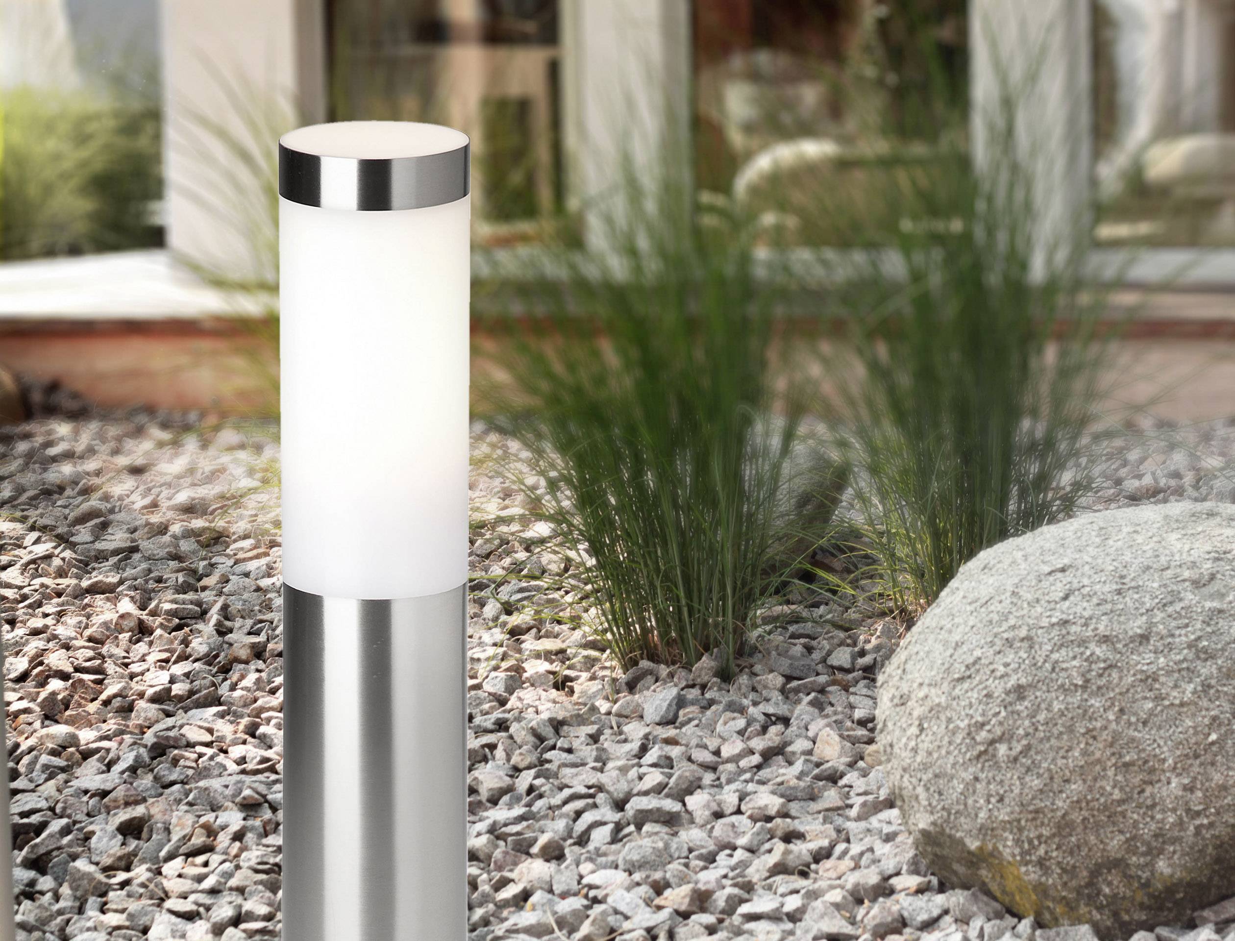 Eine moderne Außenleuchte aus Metall und Glas steht in einem Kiesgarten neben einem großen Stein und Gräsern.