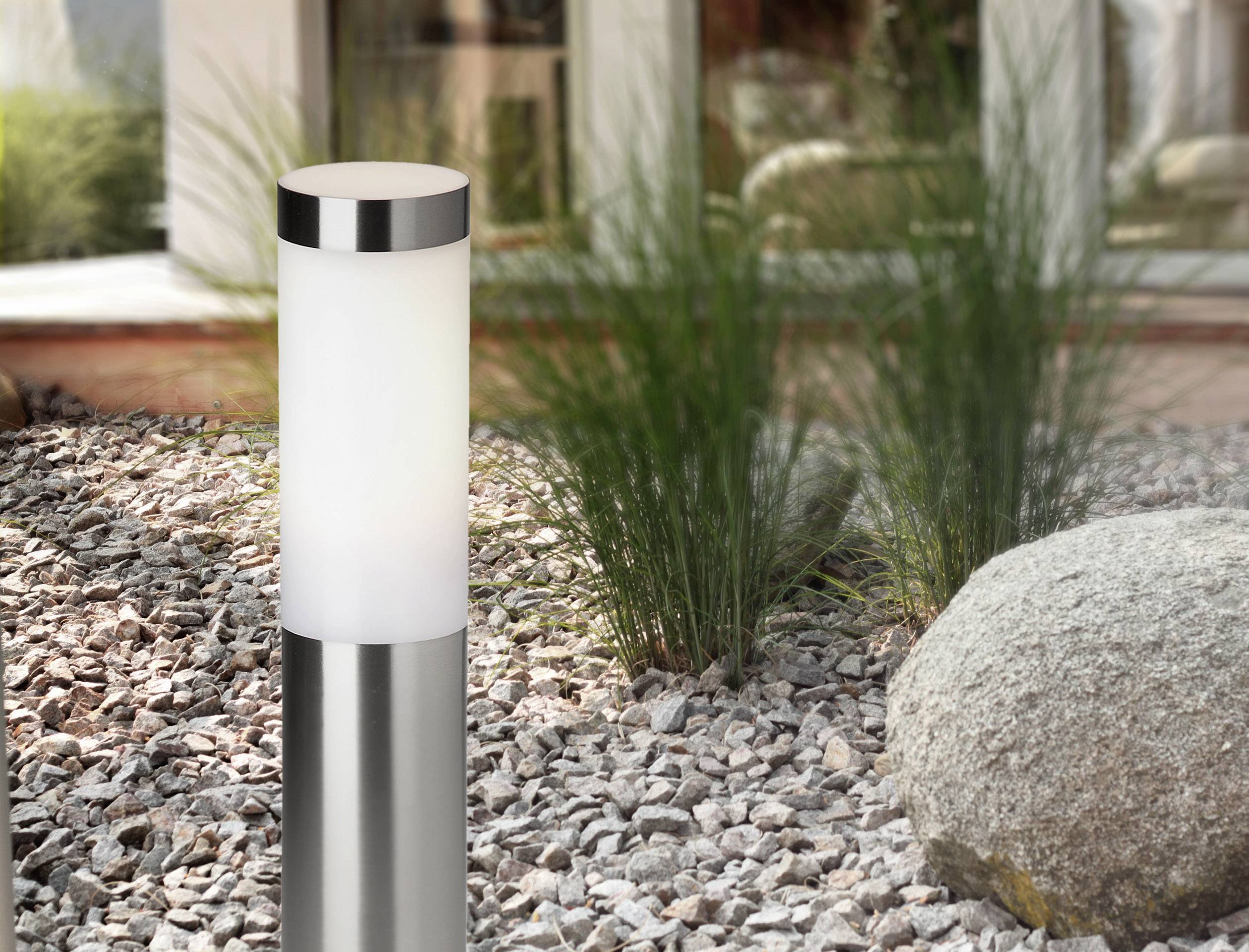 Eine moderne Gartenleuchte aus Edelstahl steht auf einem Kiesboden, umgeben von grünen Gräsern und einem großen Stein. Im Hintergrund ist ein Gebäude zu sehen.