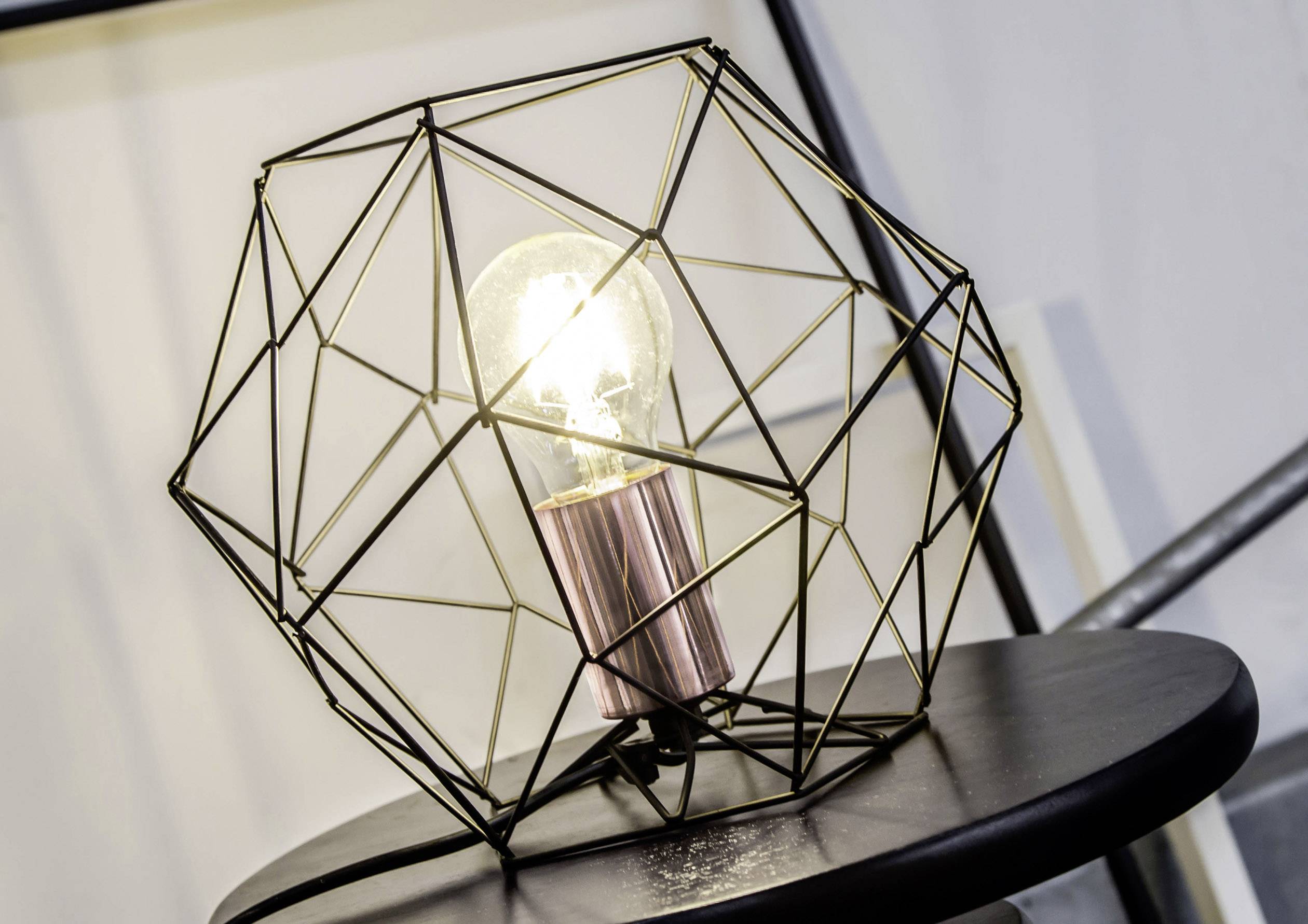 Eine Glühbirne in einem modernen, geometrischen Metallrahmen steht auf einem Tisch. Die Lampe leuchtet, was auf Verwendung hinweist.