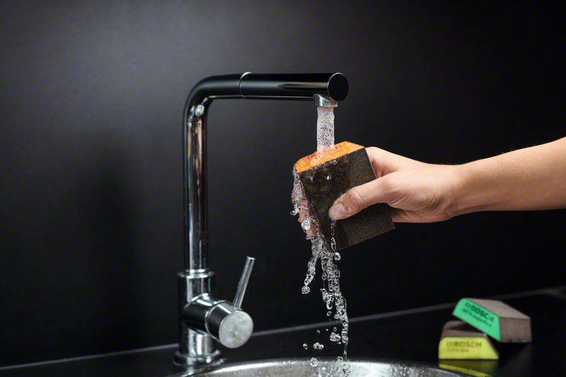 Eine Hand hält einen Schwamm unter fließendes Wasser aus einem modernen Wasserhahn in einer Küche.