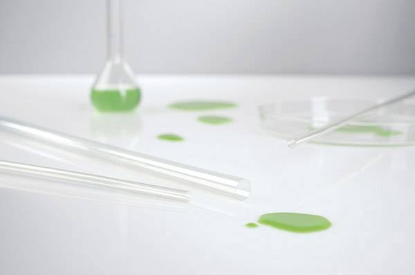 Ein umgestürztes Reagenzglas mit grüner Flüssigkeit auf einem Labortisch, weitere Laborgeräte im Hintergrund.