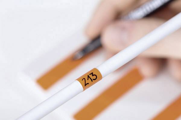 Weiße Messsonde mit der Aufschrift '213' im Vordergrund. Im Hintergrund schreibt eine Hand mit einem Stift auf ein Papier mit orangefarbenen Streifen.