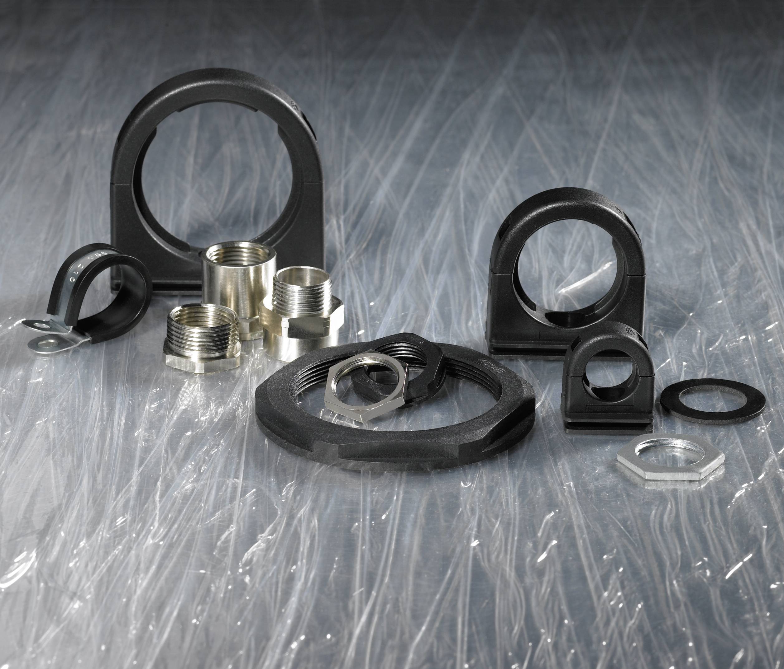 Schwarze Kunststoffklemmen und Metallverschraubungen auf einer transparenten, strukturierten Oberfläche. Zubehör für Rohr- oder Kabelmontage.