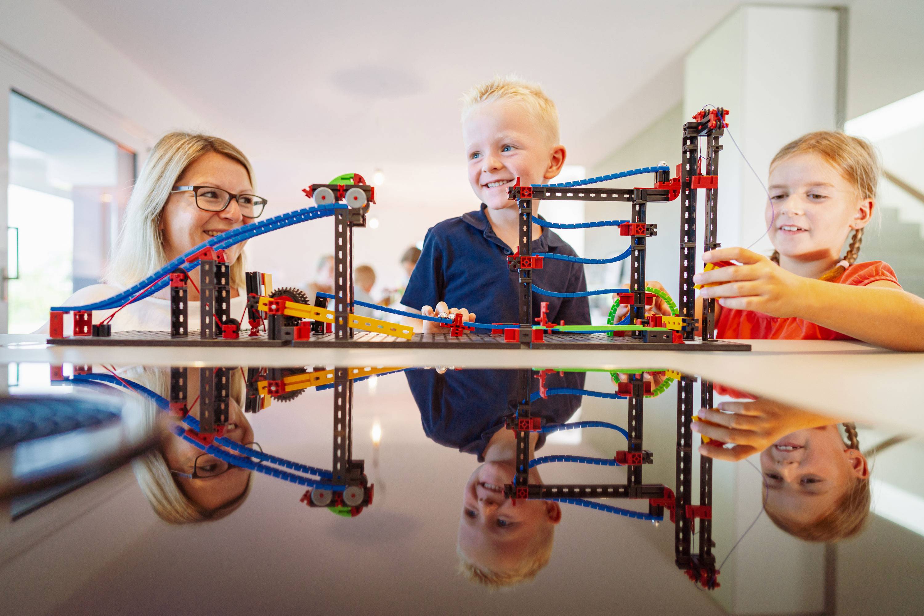 Drei Kinder spielen fröhlich mit einer bunten Lego-Achterbahn auf einem Tisch. Sie lachen und zeigen Freude am gemeinsamen Spiel.