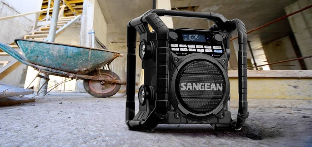 Tragbares Baustellenradio auf einem Baustellenboden, neben einer Schubkarre. Robust und funktional für Musik oder Nachrichtenempfang.