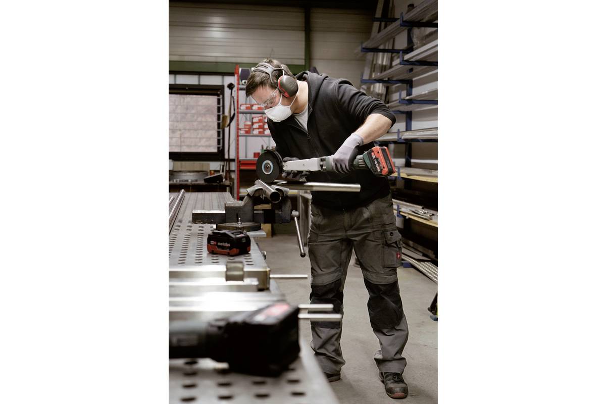 Arbeiter in Schutzkleidung schleift Metall in einer Werkstatt. Er benutzt eine elektrische Schleifmaschine auf einem Arbeitstisch.