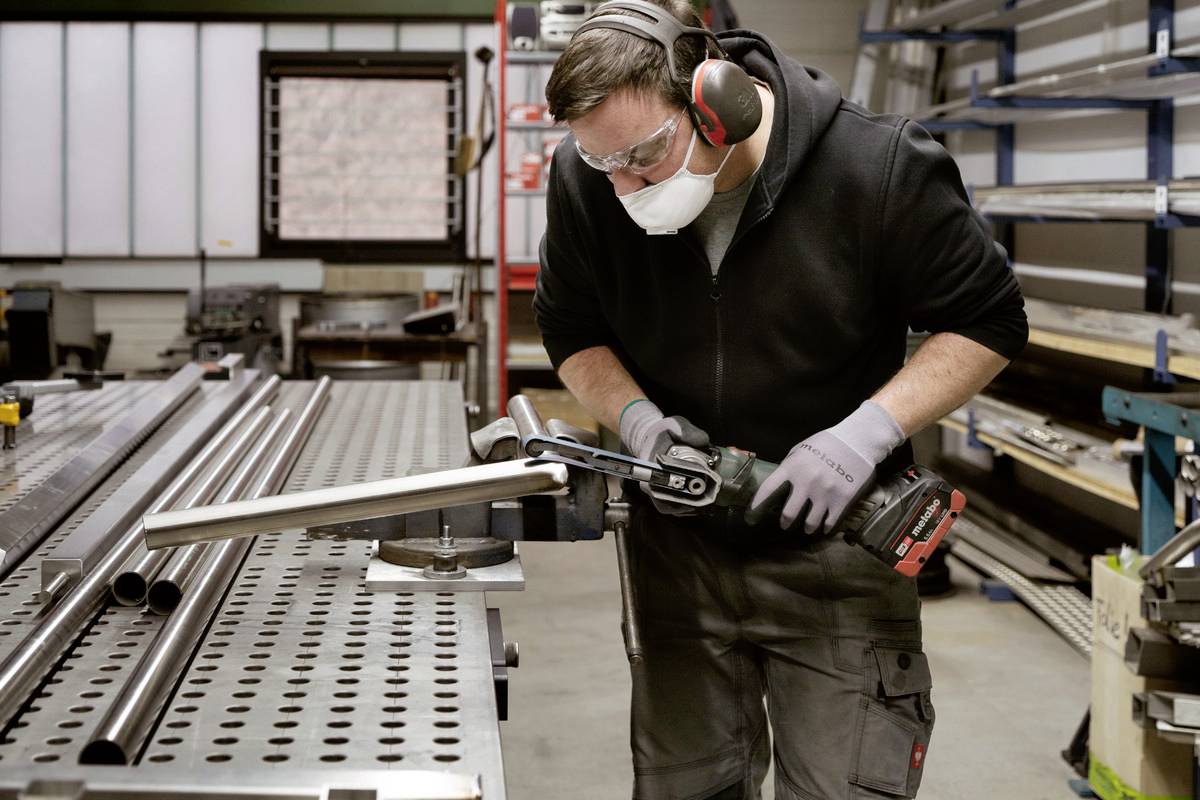Eine Person in Schutzkleidung schleift Metall in einer Werkstatt mit einem Winkelschleifer. Im Hintergrund sind Regale und Werkzeuge.