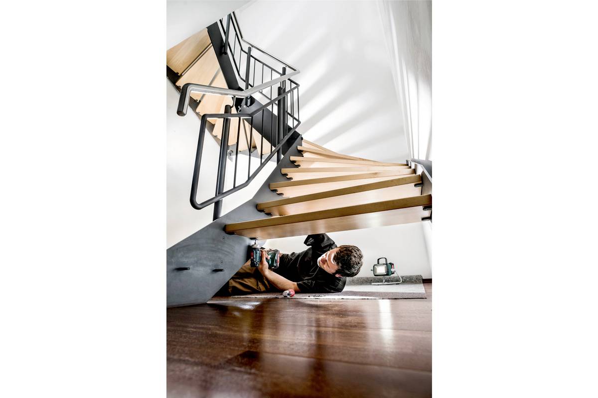 Ein Mann arbeitet mit einem Werkzeug unter einer modernen Holztreppe mit Metallgeländer. Im Hintergrund sind Werkzeugkoffer sichtbar.