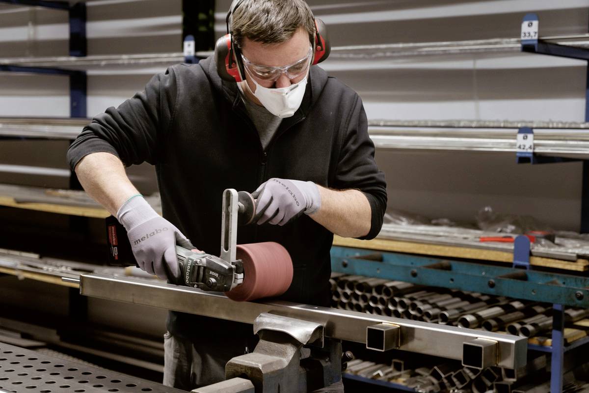 Ein Mann schleift mit einer Schleifmaschine ein Metallstück in einer Fabrik. Er trägt Schutzbrille, Gehörschutz und eine Maske.